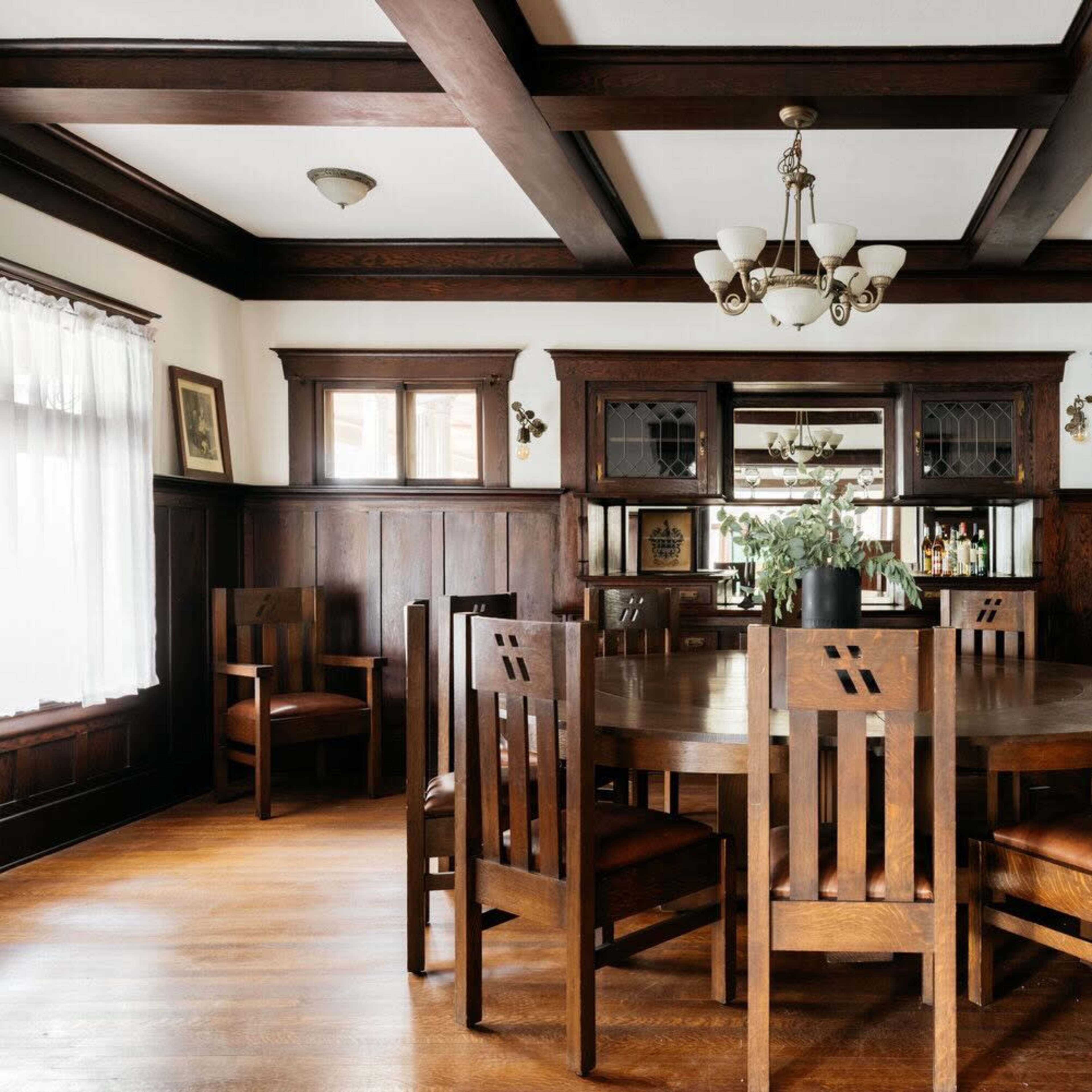A wooden dining room with a large circular table surrounded by eight chairs, framed by dark wood paneling and a built-in cabinet.