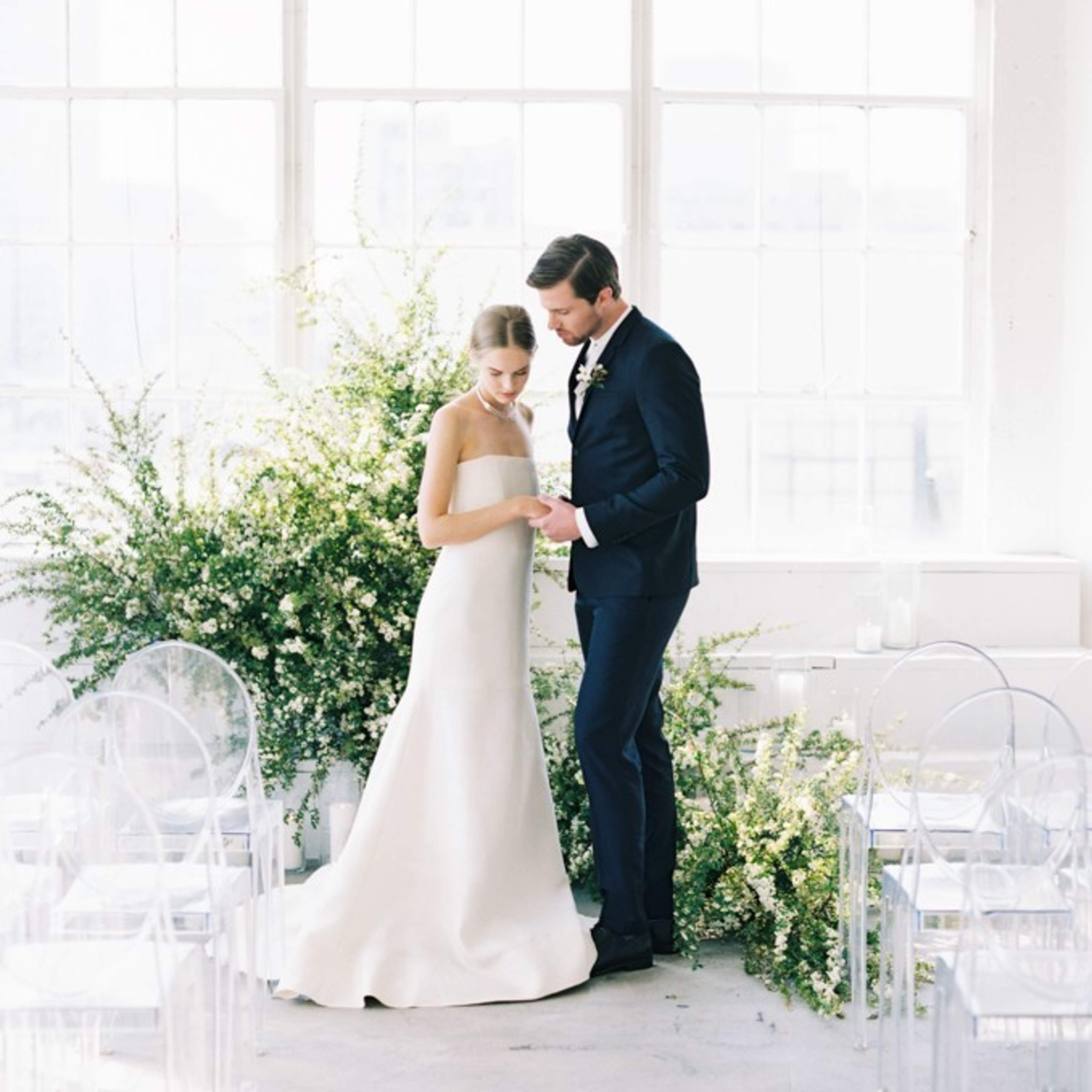A bride and groom stand together in a sunlit room adorned with greenery, preparing for their wedding ceremony.