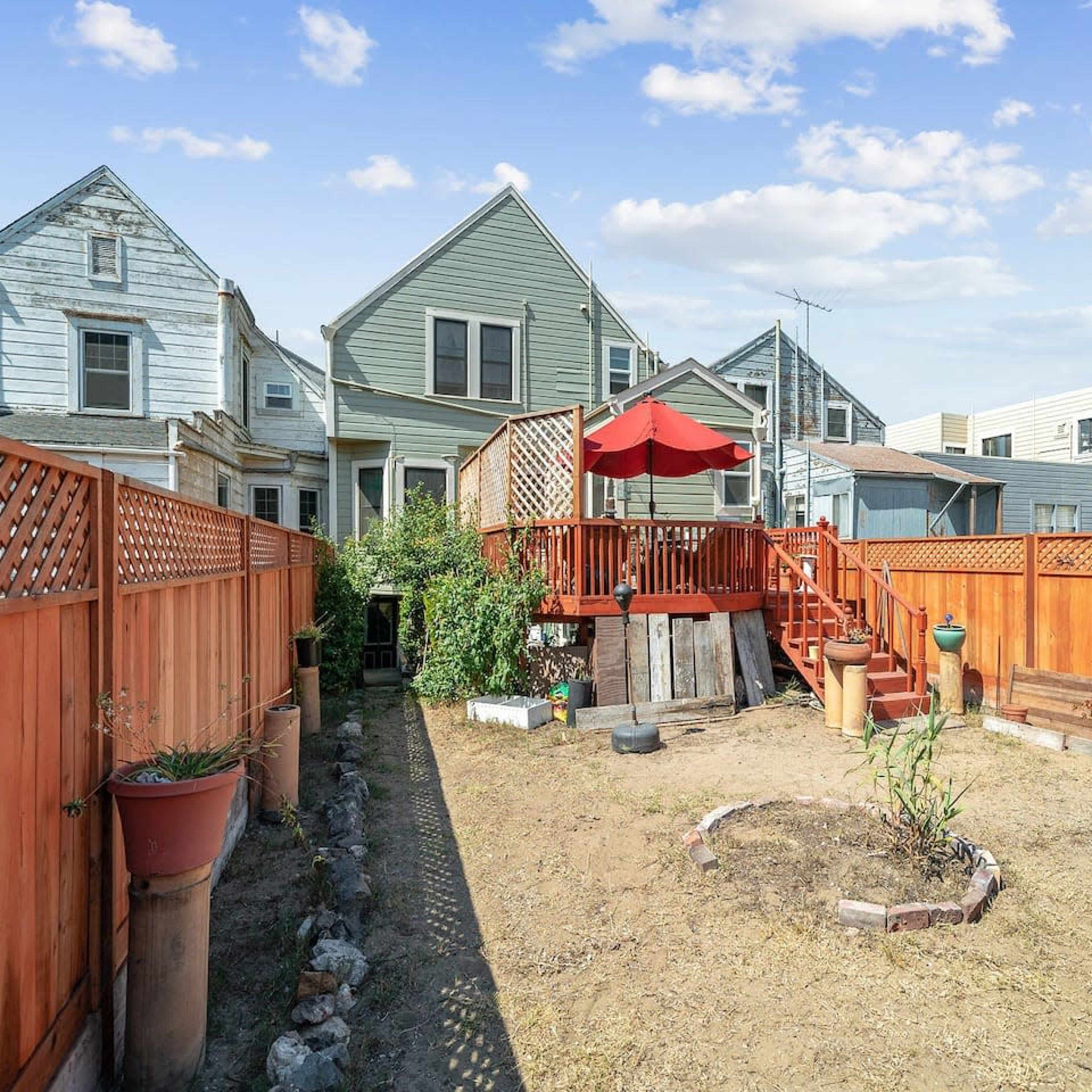 A fenced backyard with a wooden deck, an umbrella, and a small garden area surrounded by residential buildings.