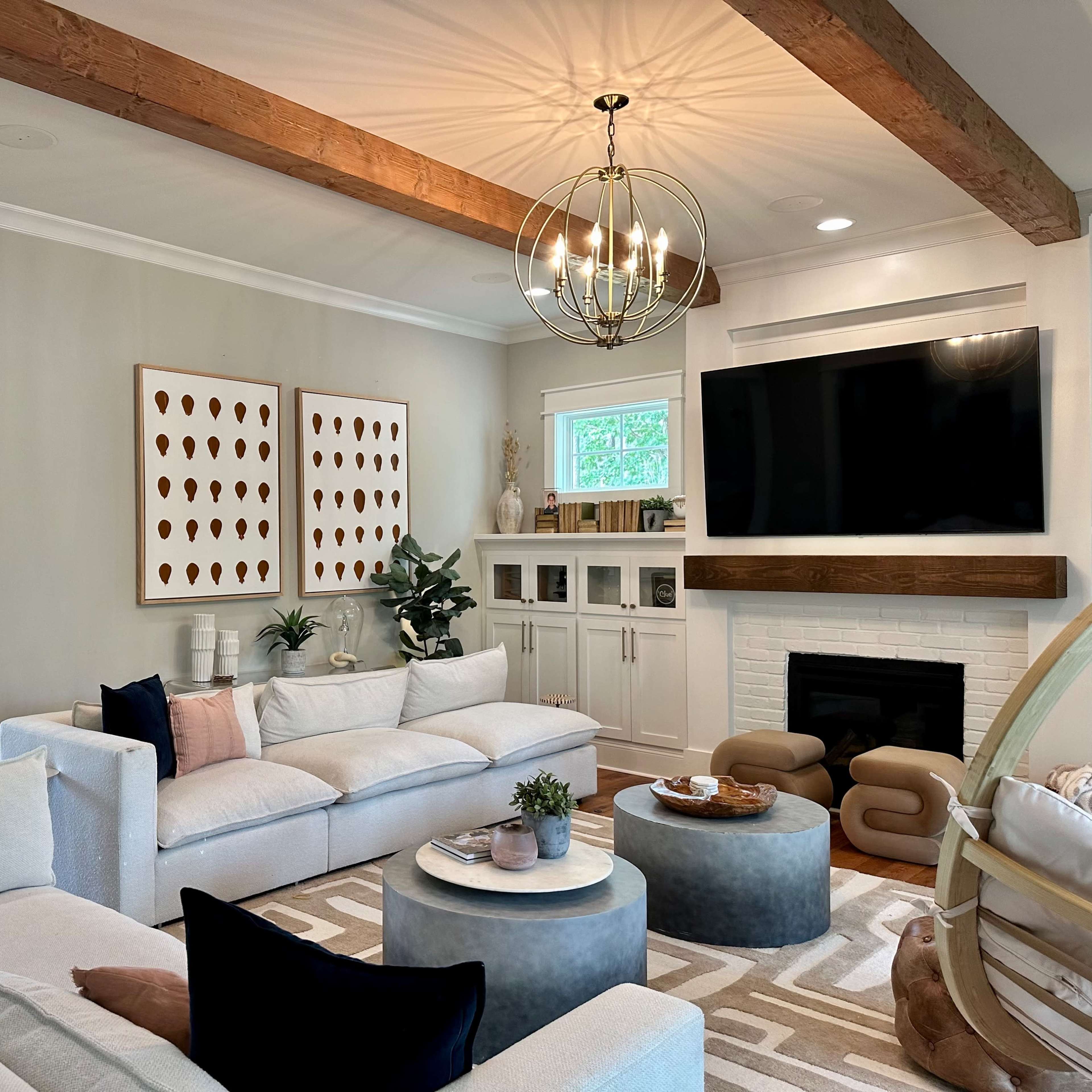 The living room features a light-colored sectional sofa, a round coffee table, and a mounted flat-screen TV above a white fireplace, with wooden beams and decorative artwork on the walls.
