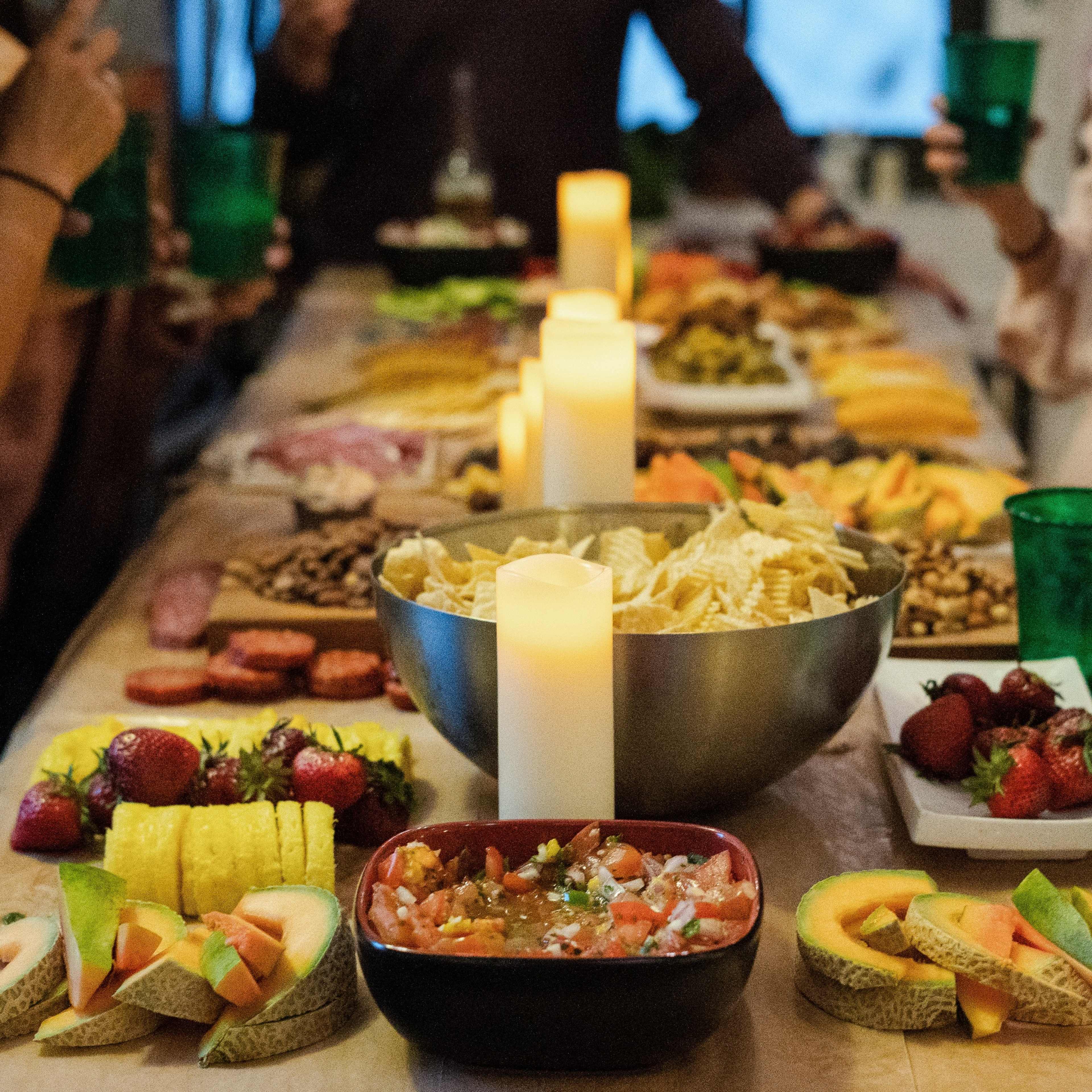 A long table is adorned with various dishes, including fruits, snacks, and dips, surrounded by guests holding drinks.