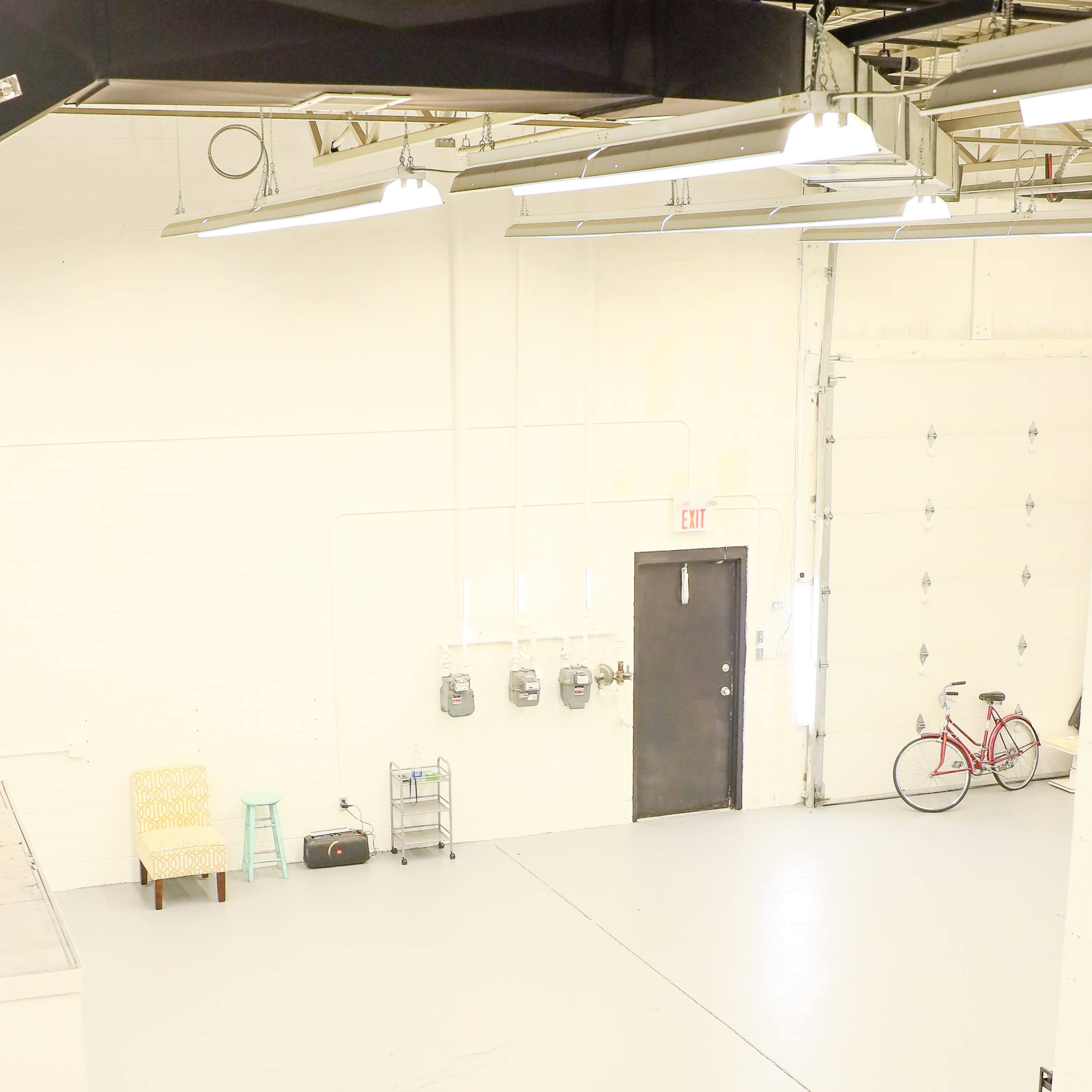 A spacious industrial room with concrete flooring, a closed door, a bicycle, and a few pieces of furniture along the walls.