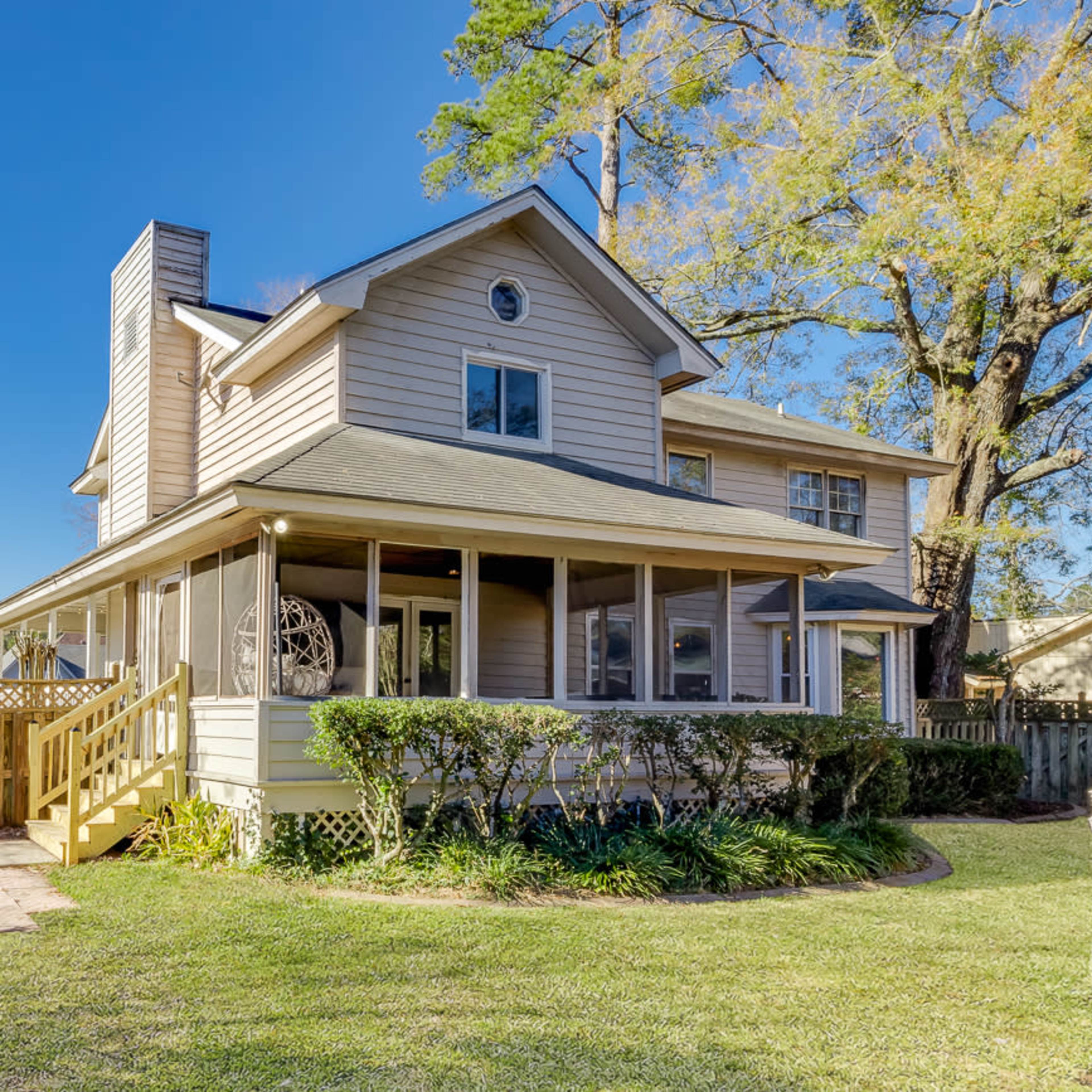 A two-story house with a screened porch and wooden stairs is surrounded by greenery and a fenced backyard.