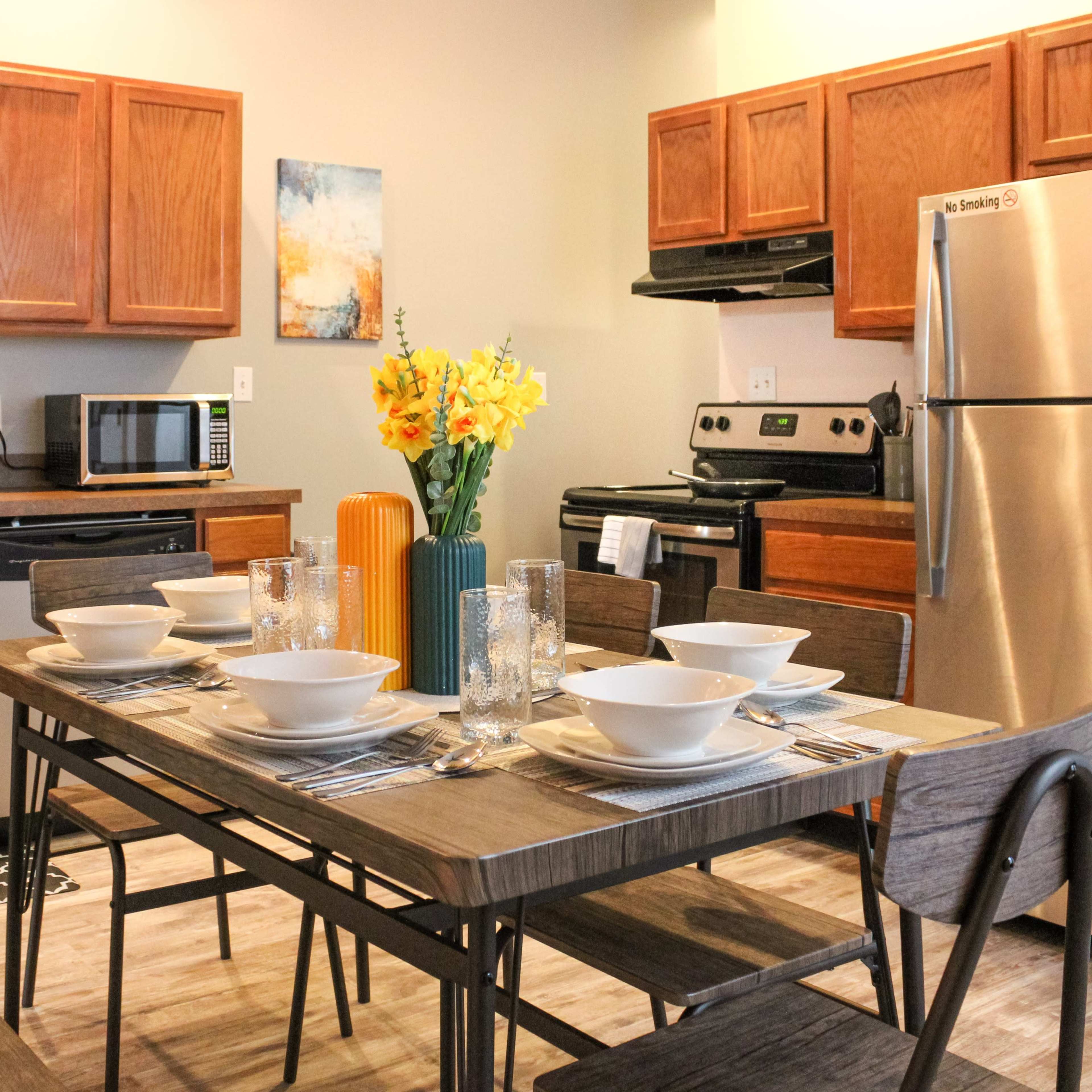 A modern kitchen features wooden cabinets, stainless steel appliances, and a dining table set for six with bowls and glasses, complemented by a floral centerpiece.