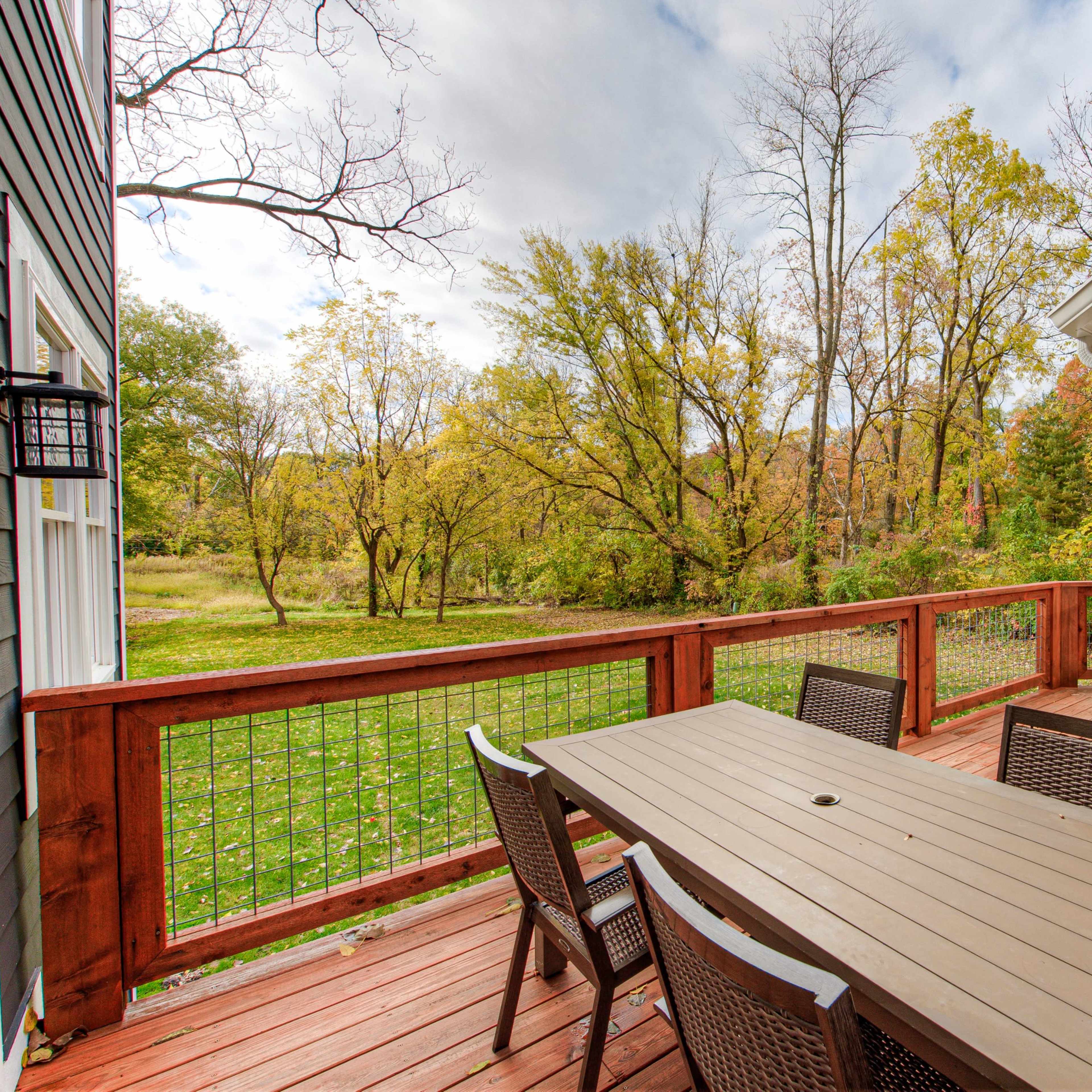 A wooden deck features a table and chairs, overlooking a grassy area with trees in the background.