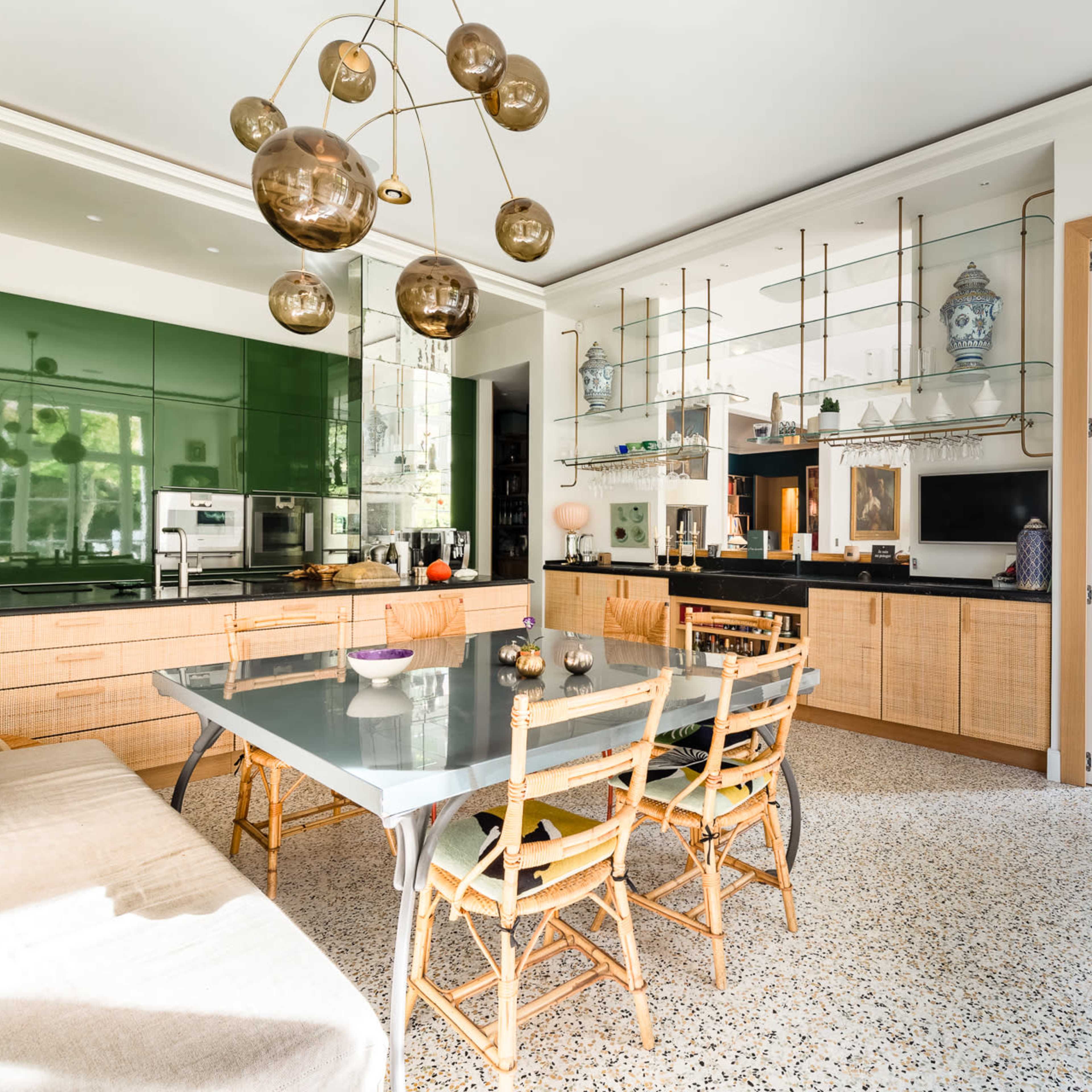 A modern dining area features a long table surrounded by wooden chairs, with a green kitchen backdrop and decorative shelves above.