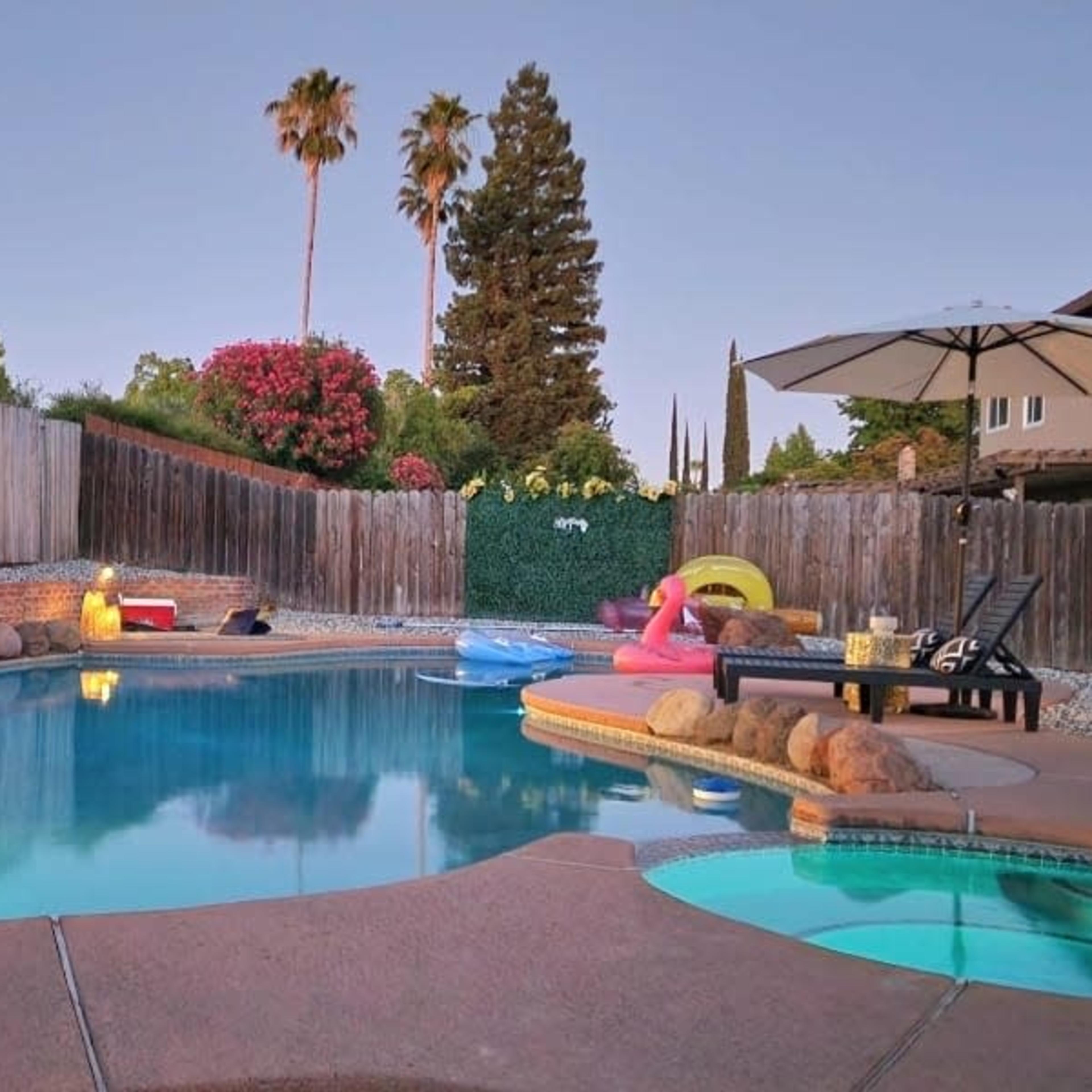 A backyard pool surrounded by trees and decorative lights, with lounge chairs and floating toys on the water.