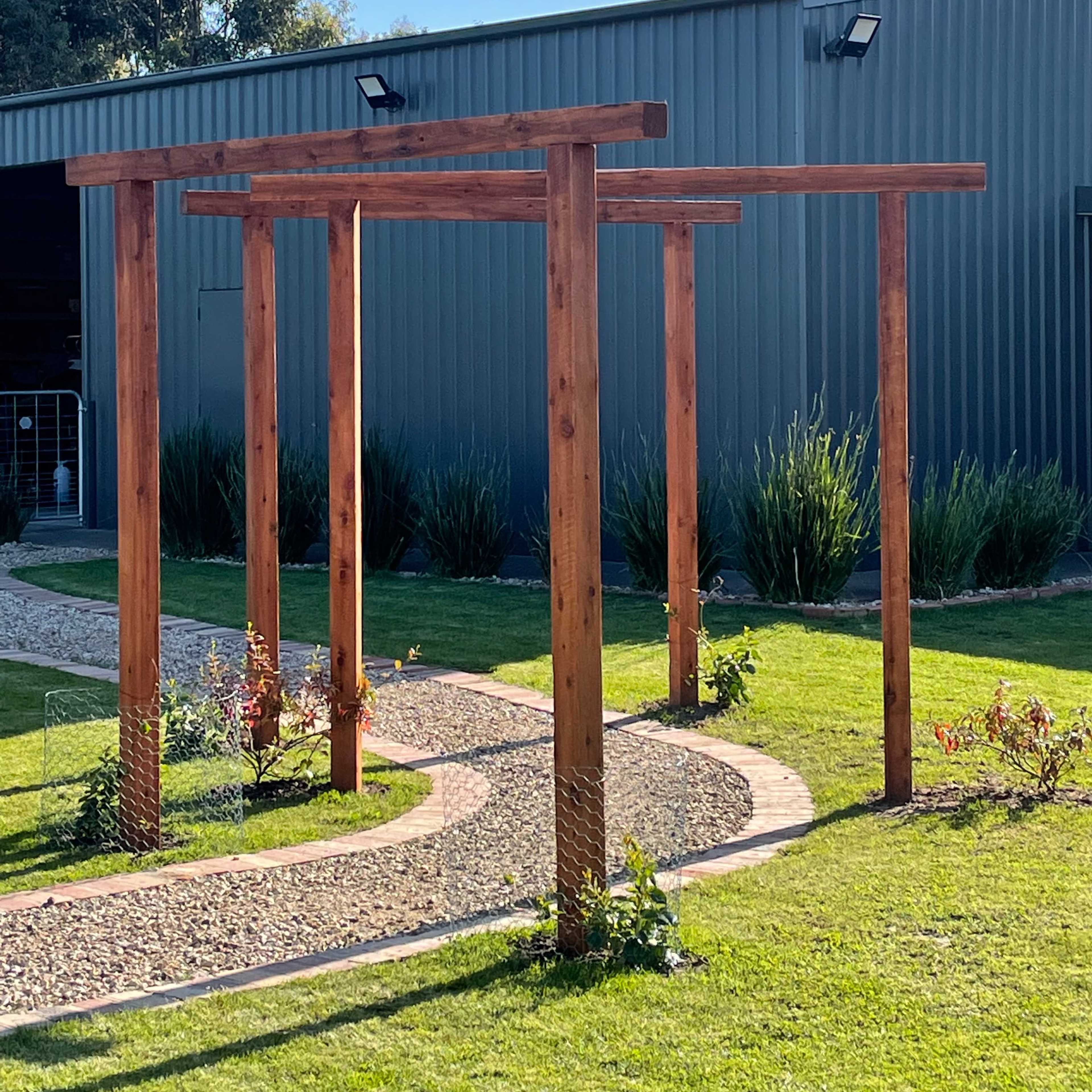 A wooden pergola structure with four vertical posts, set on a lawn beside a gravel path and a grey metal building.