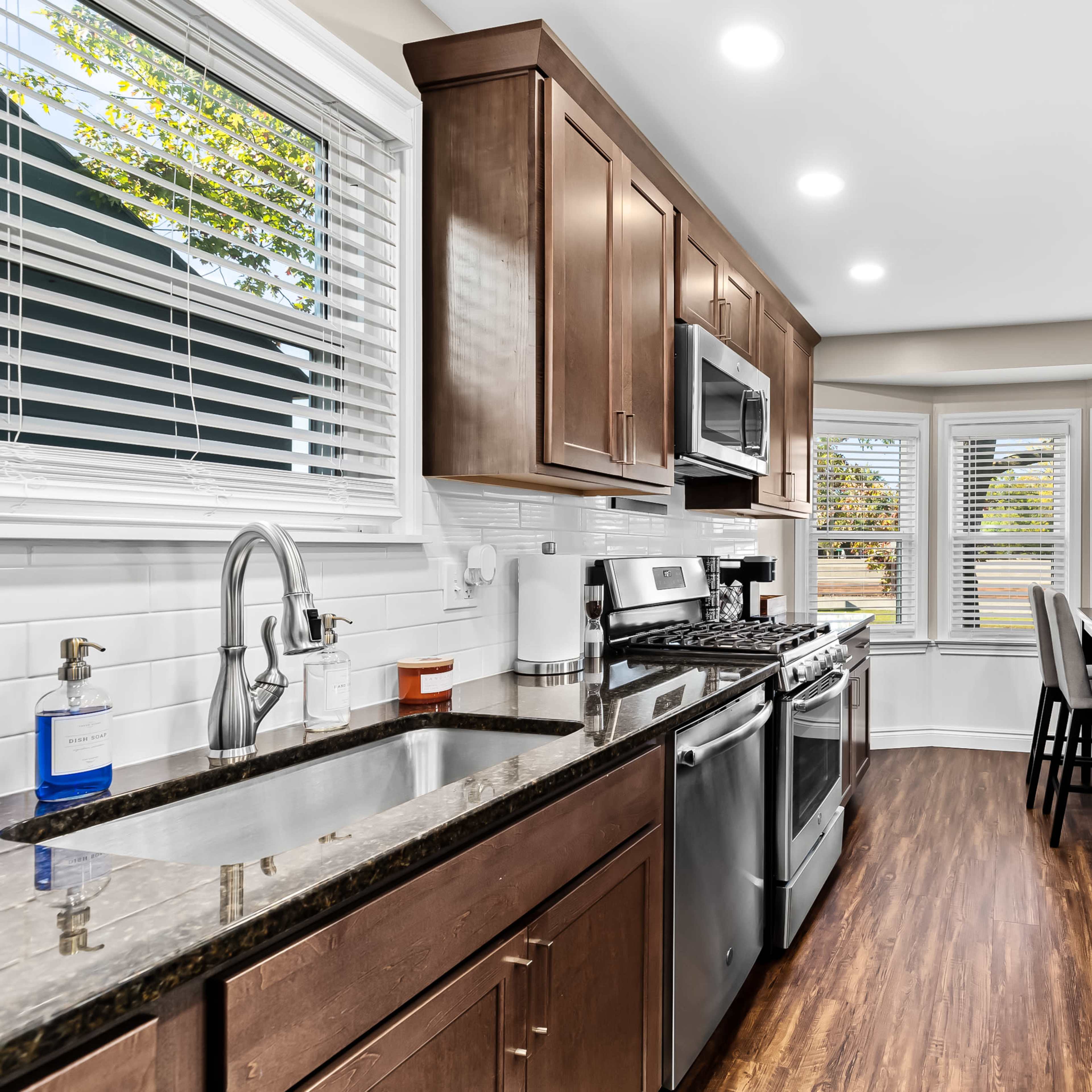 A modern kitchen with dark wood cabinets, stainless steel appliances, and a light countertop, featuring a dining area with large windows.