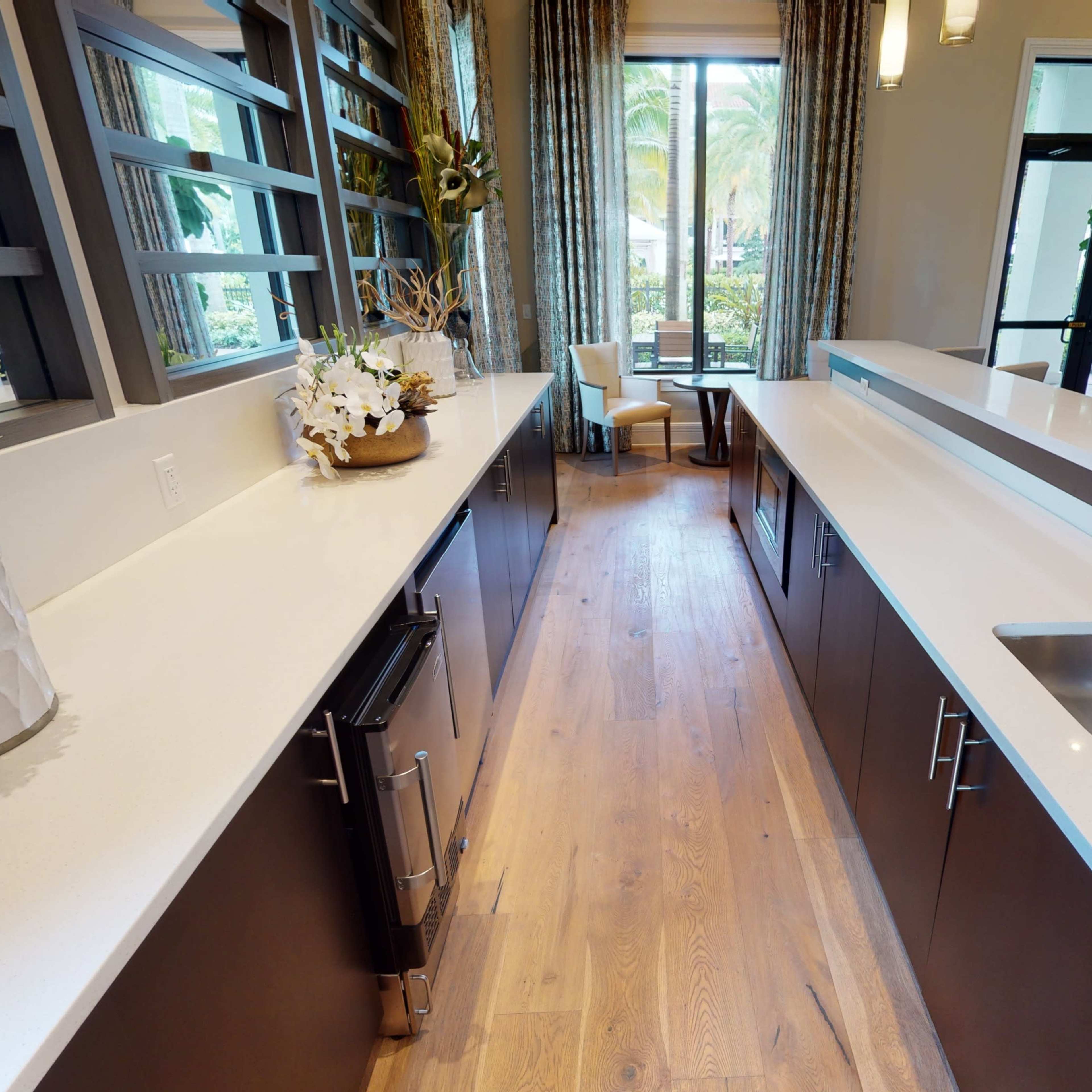 A modern kitchen with two parallel countertops, a sink on the right, and a view of a seating area and windows in the background.