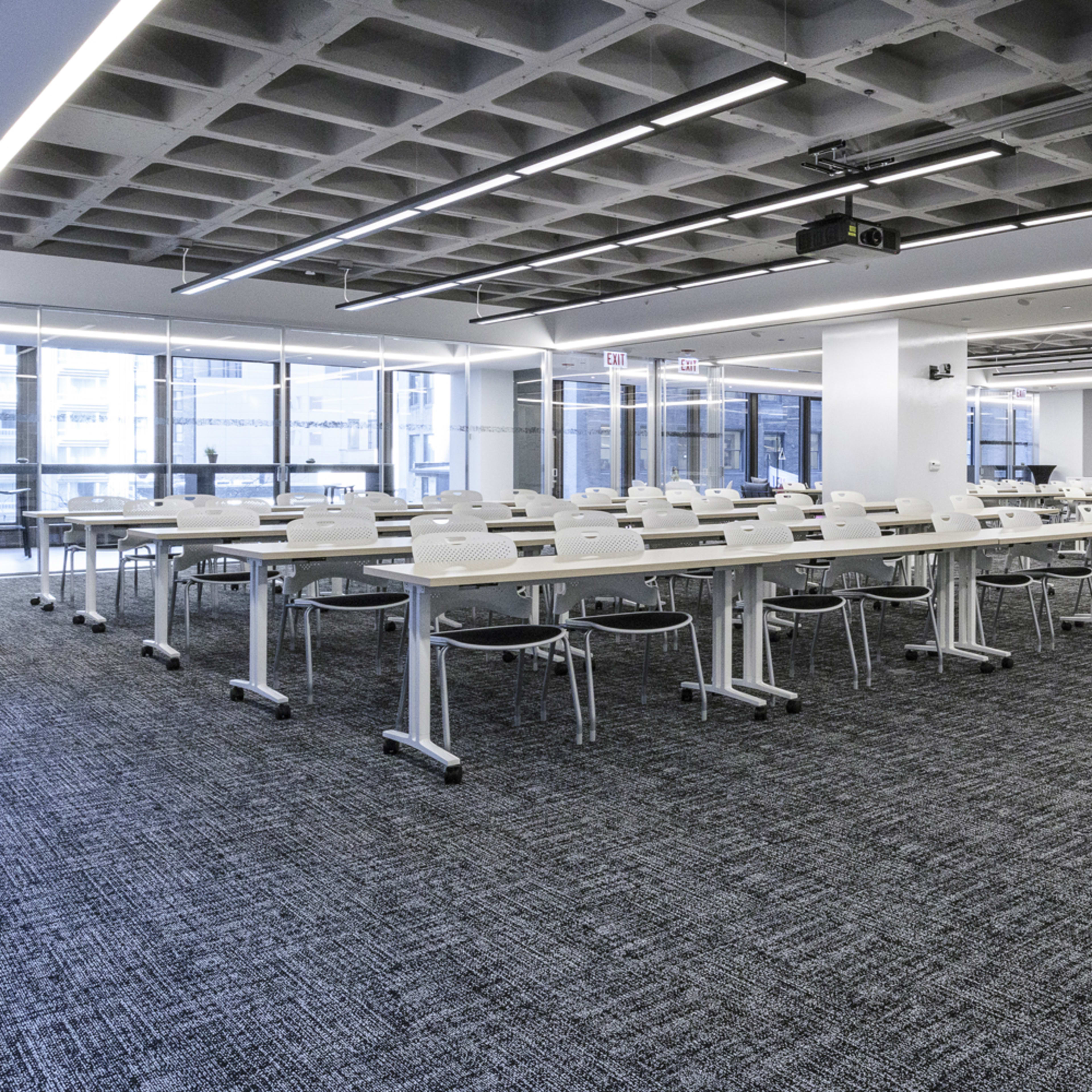 A modern classroom features numerous white tables and chairs arranged in a grid, with large windows allowing natural light and a projector mounted on the ceiling.