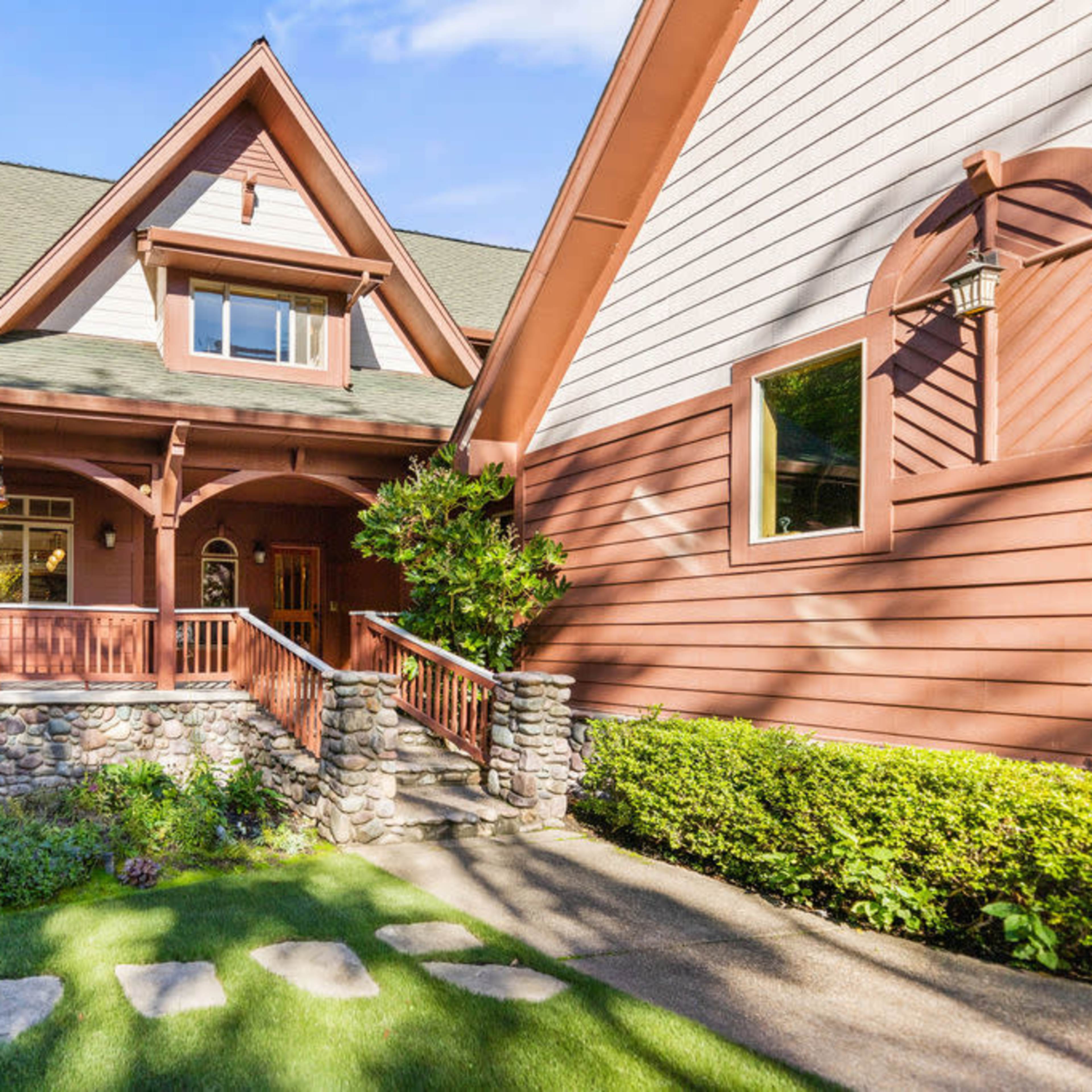 A two-story house with a wooden porch, stone accents, and a sloped roof surrounded by green landscaping.