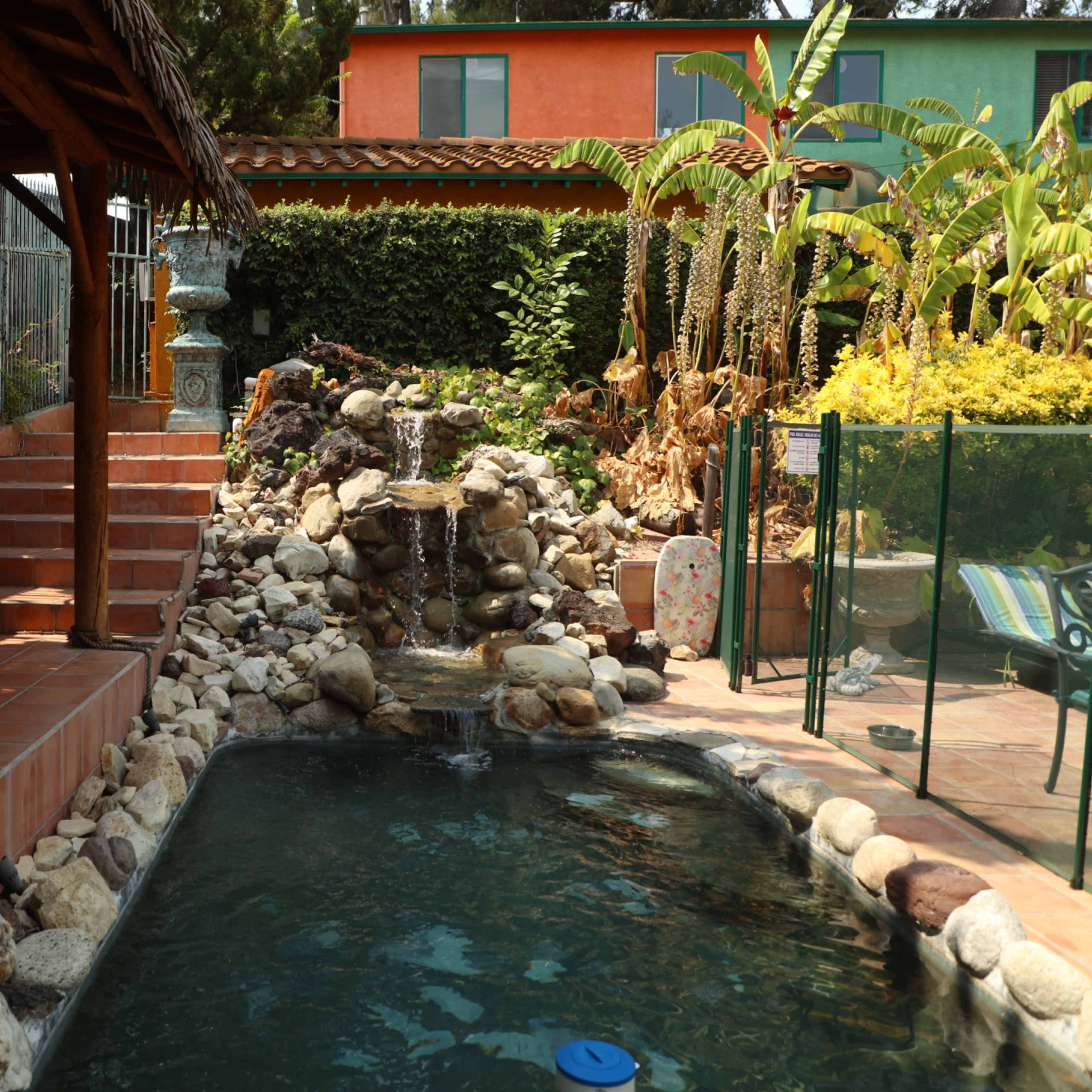 A landscaped backyard featuring a stone fountain beside a swimming pool with surrounding stones and a thatched roof gazebo.