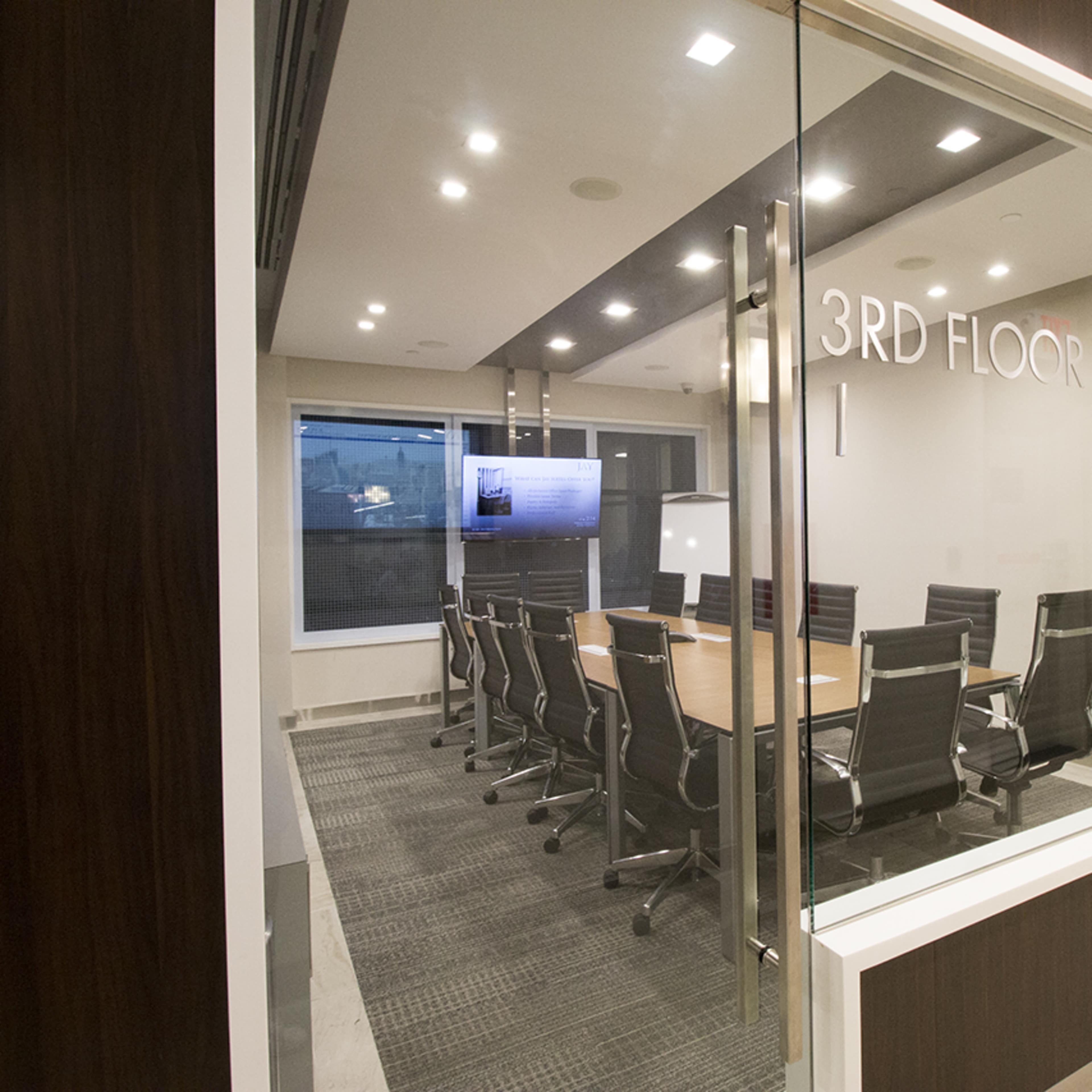 A modern conference room with a long table surrounded by black chairs, visible through glass walls.