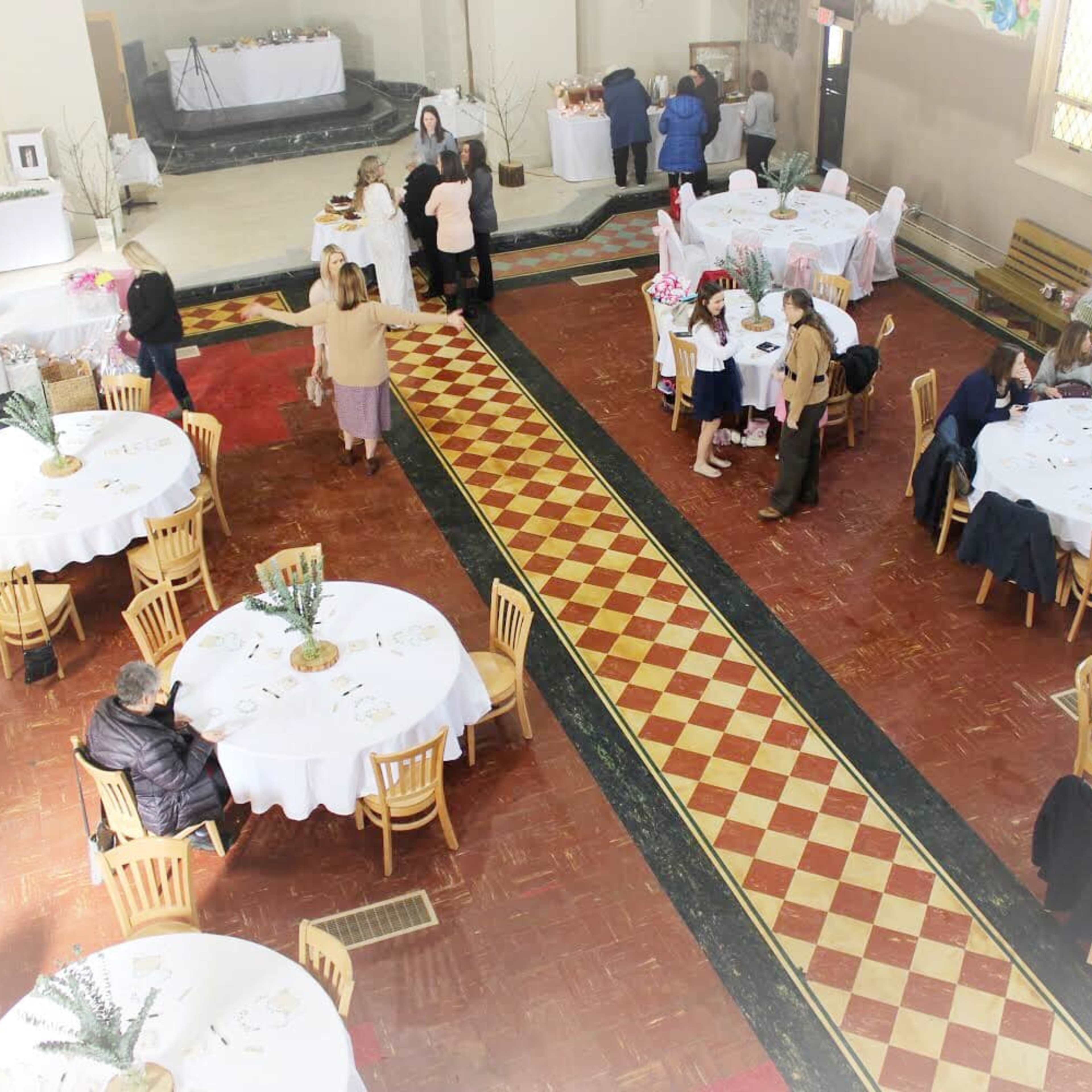 A spacious event hall is set up with round tables covered in white tablecloths, with guests mingling and a buffet area in the background.