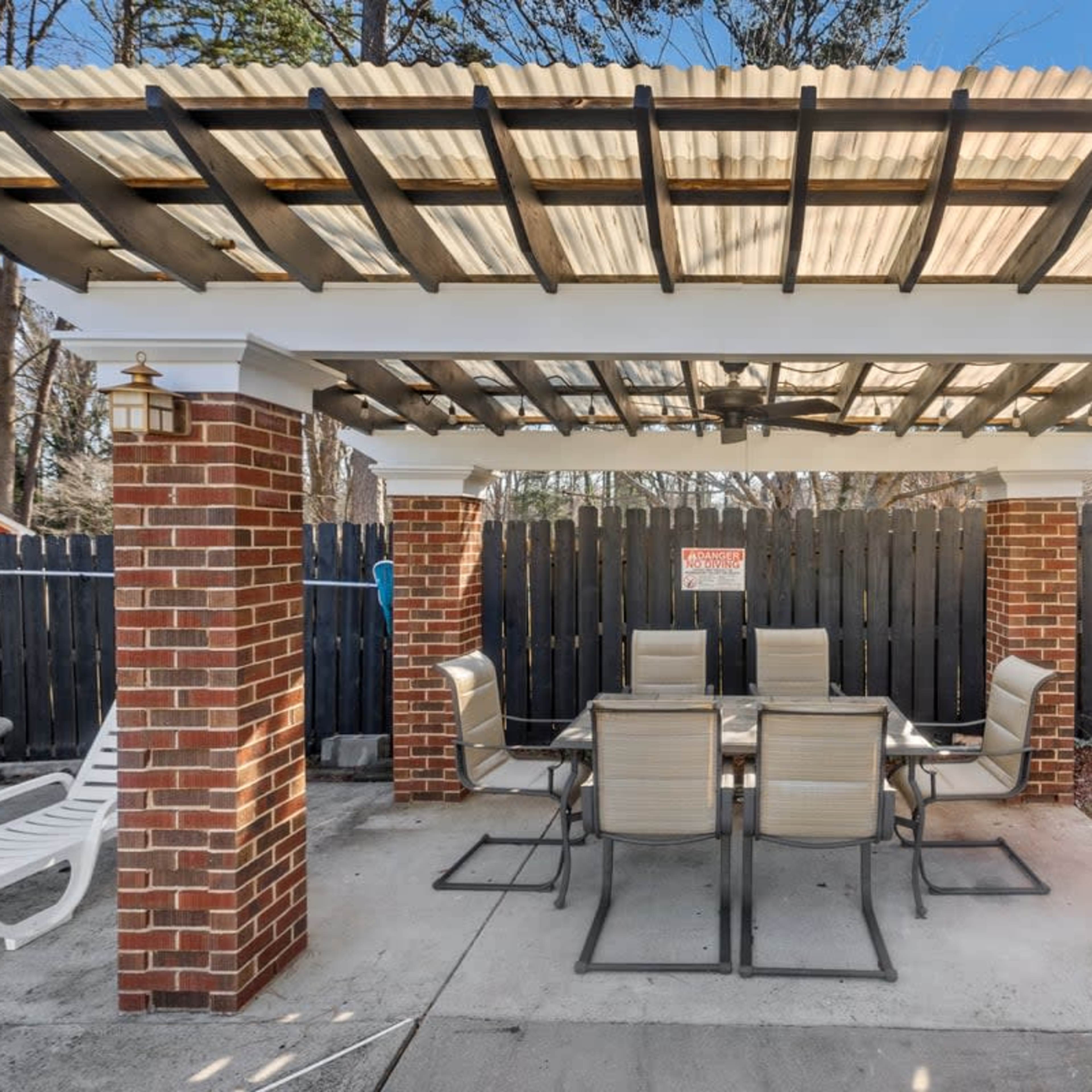 A brick patio structure with a roof, featuring a dining table and chairs underneath, surrounded by a wooden fence.
