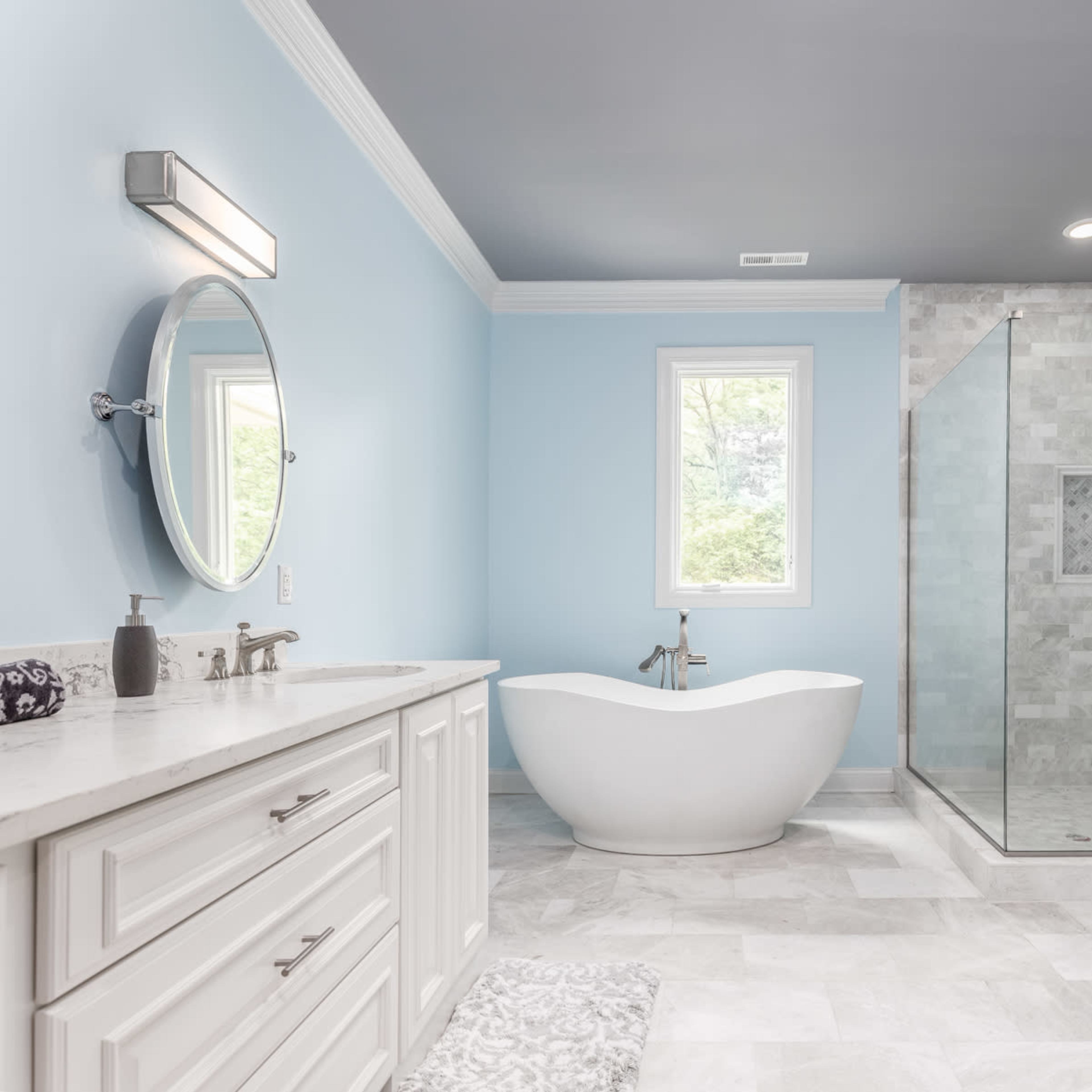 A modern bathroom featuring a glass shower, a freestanding tub, and light blue walls with white cabinetry.