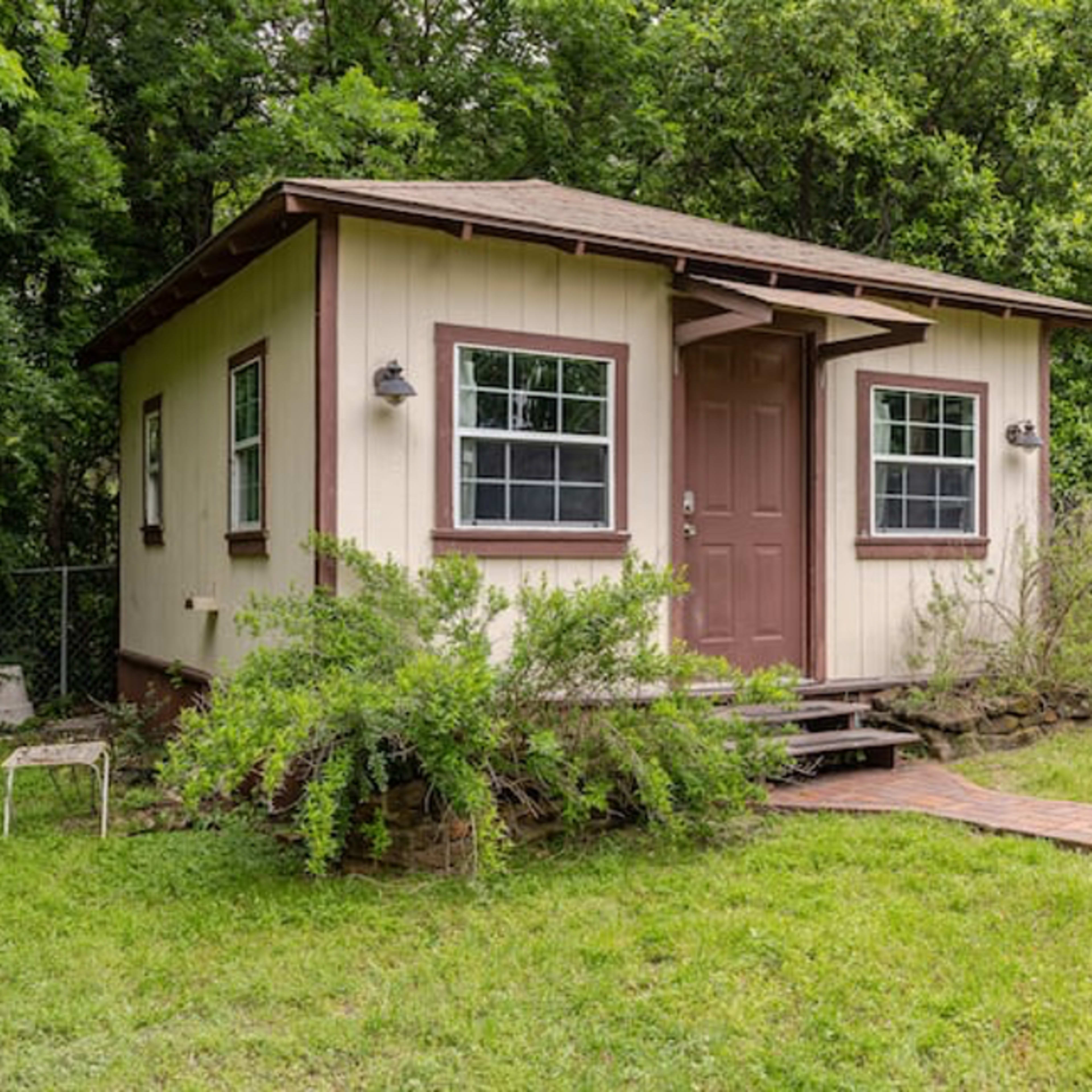 A small, single-story building with a porch sits surrounded by grass and trees.