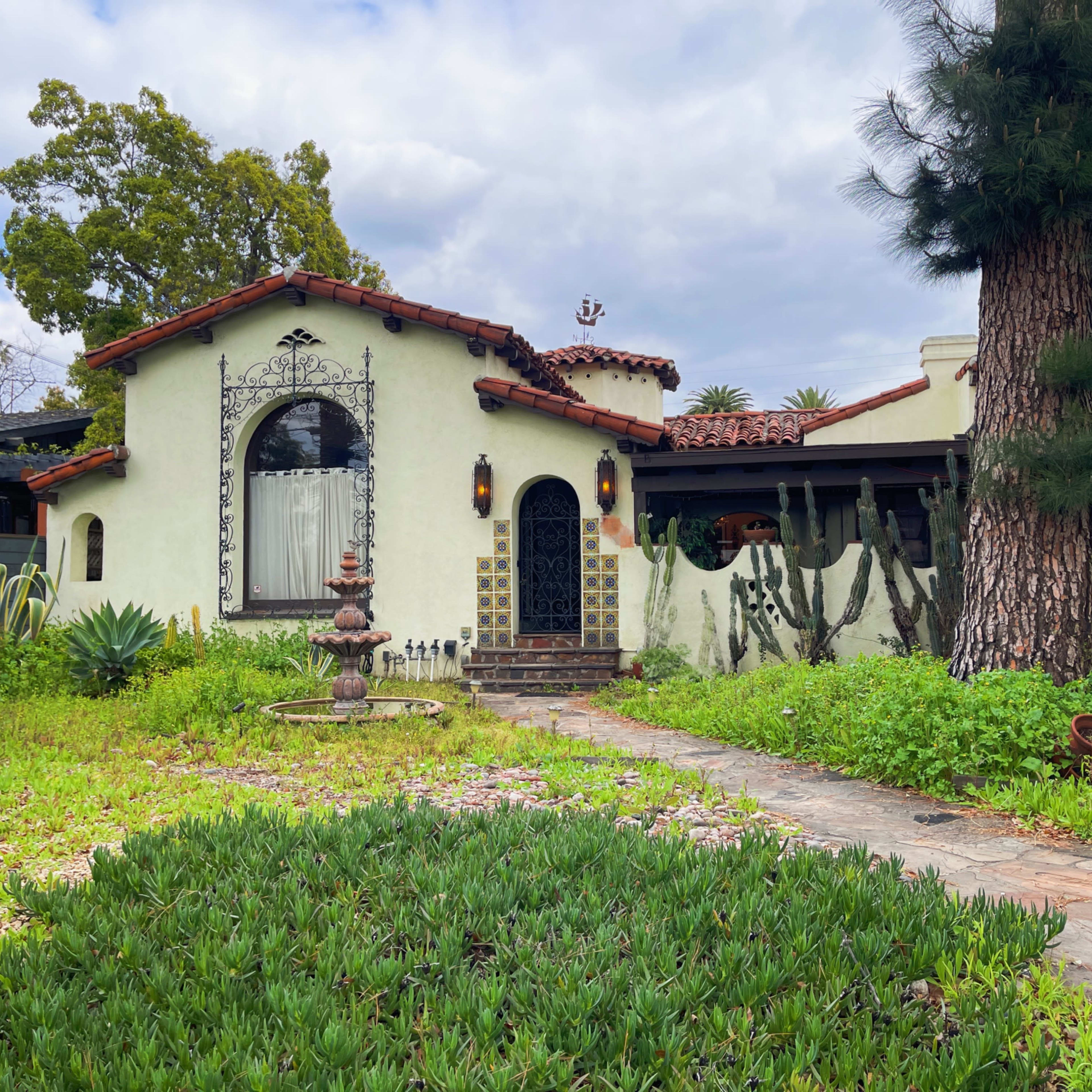A stucco house with a terracotta roof, a front garden featuring green plants, and a stone pathway leading to the entrance.