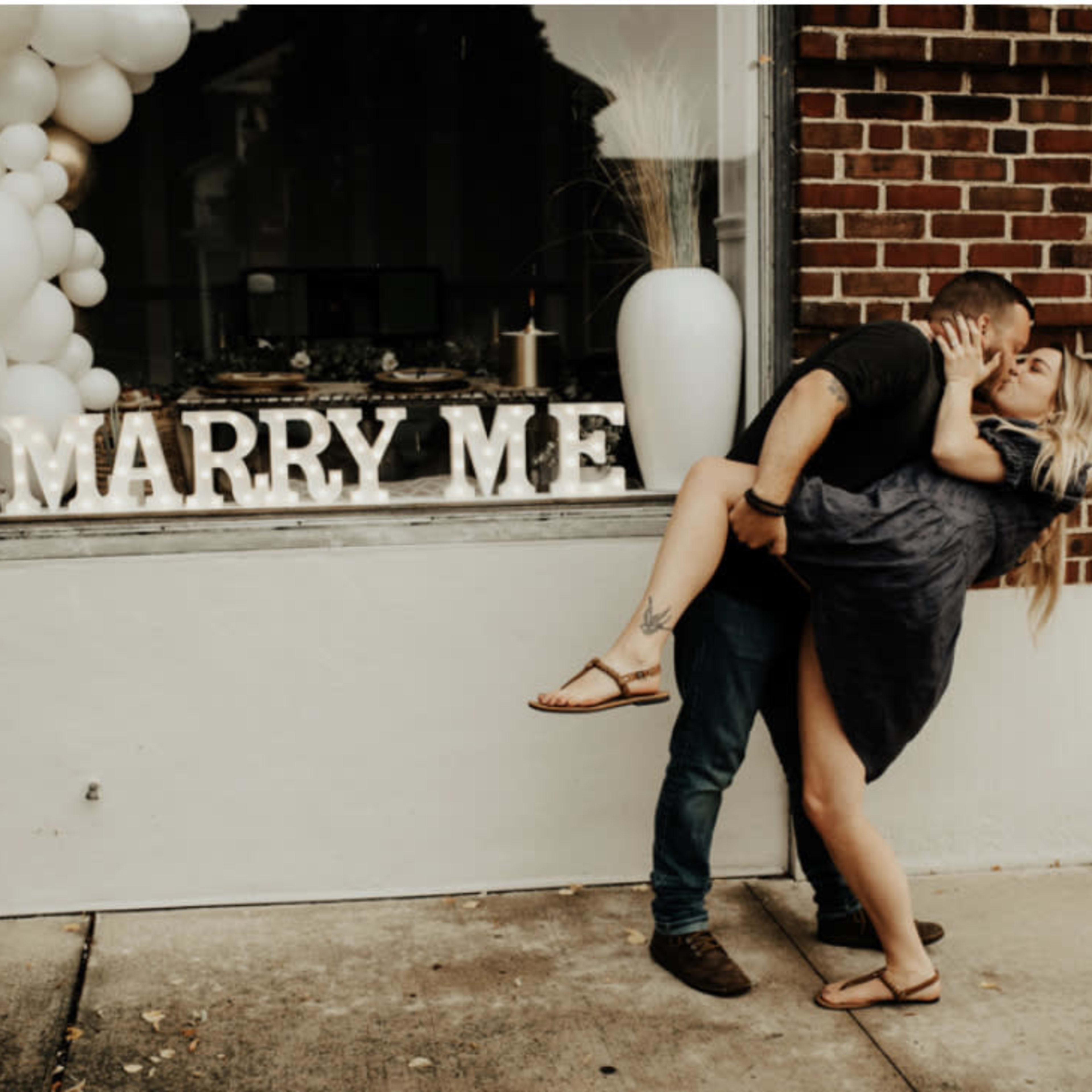 A couple embraces passionately outside a store featuring a "MARRY ME" display.
