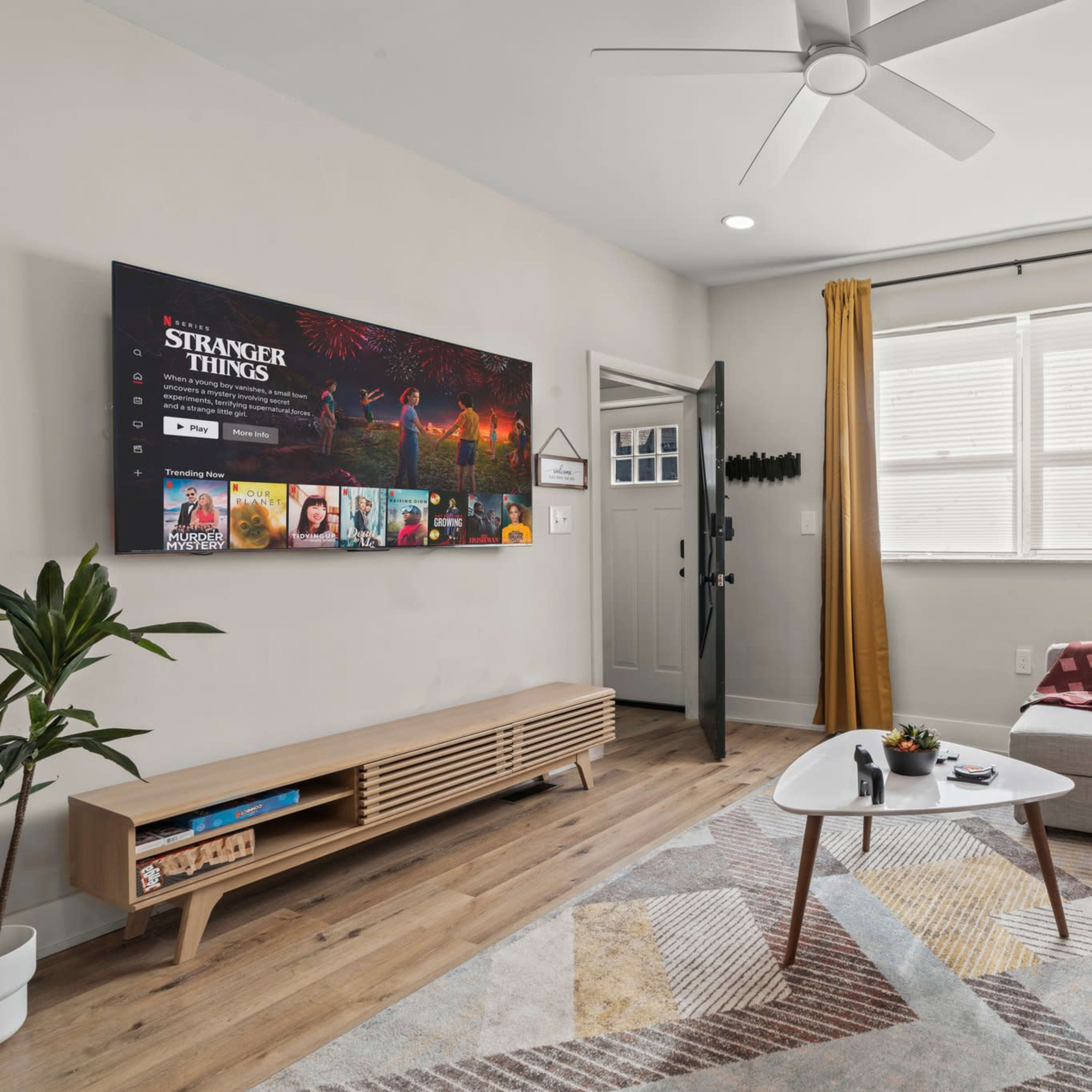 A modern living room features a large TV mounted on the wall, a gray sofa with pink cushions, a wooden coffee table, and an open door leading outside.