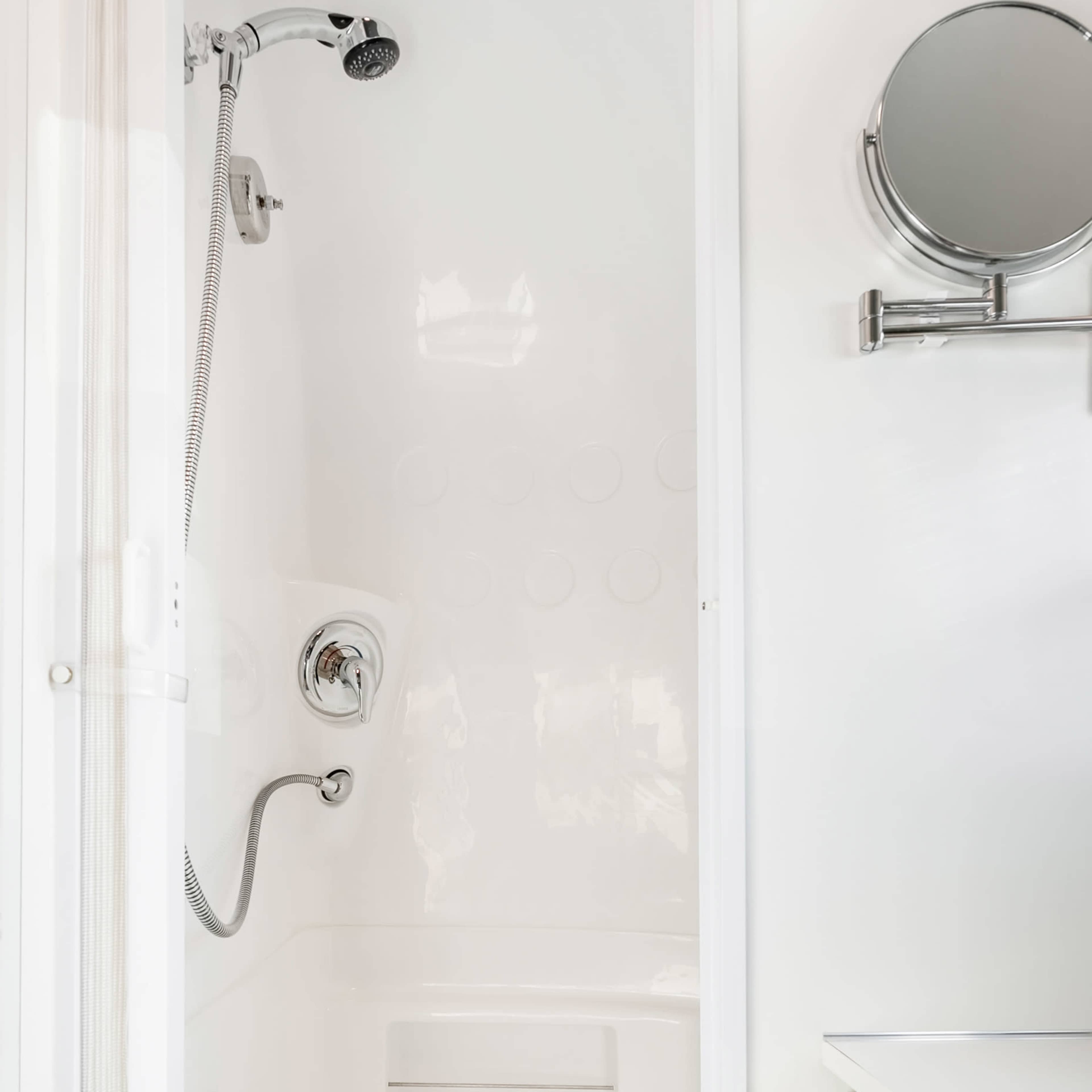 A modern shower enclosure with a white interior, featuring a handheld showerhead and a round mirror mounted on the wall.