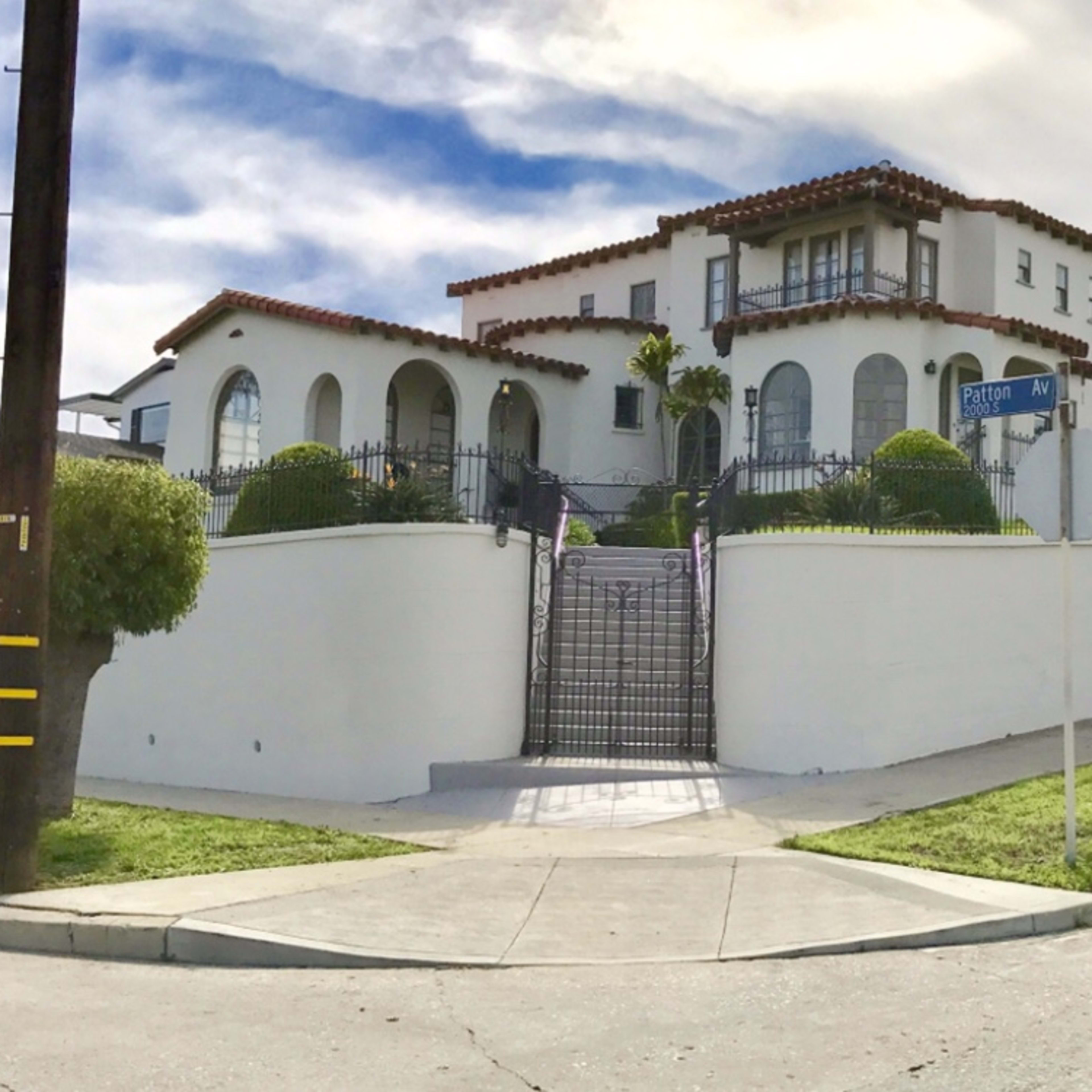 A large, two-story white house with a tiled roof sits on a landscaped corner lot, surrounded by a low white wall and ornamental trees.