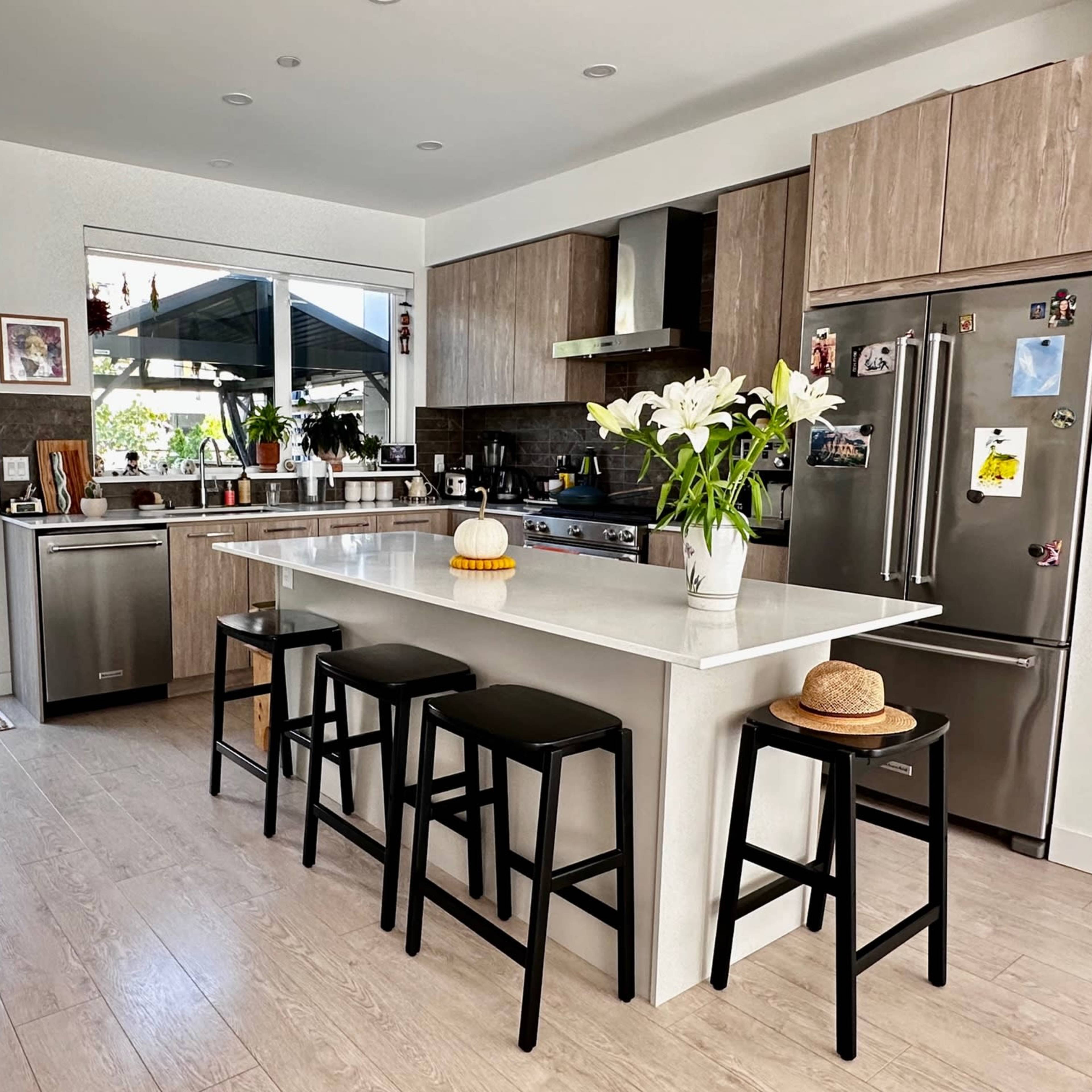 A modern kitchen features a large island with black stools, stainless steel appliances, and a view of a garden through the open door.