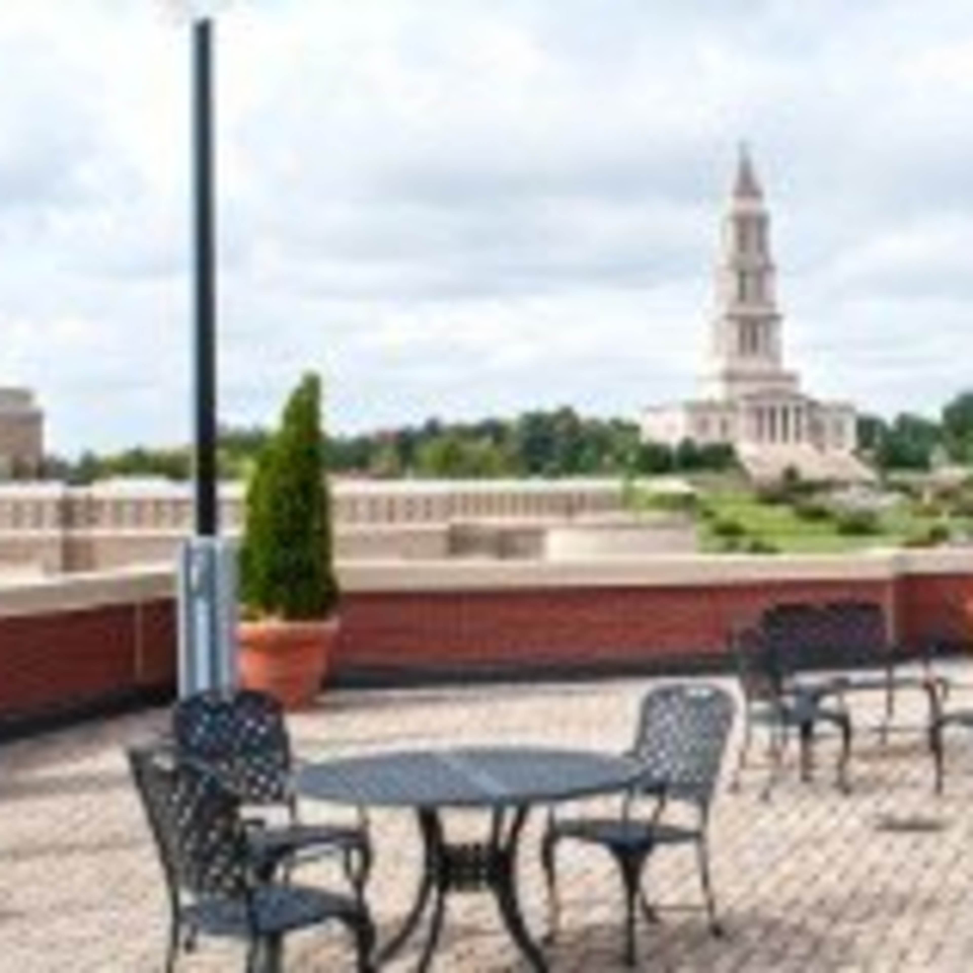 A rooftop terrace features several metal tables and chairs overlooking a landscaped area with a prominent building in the background.
