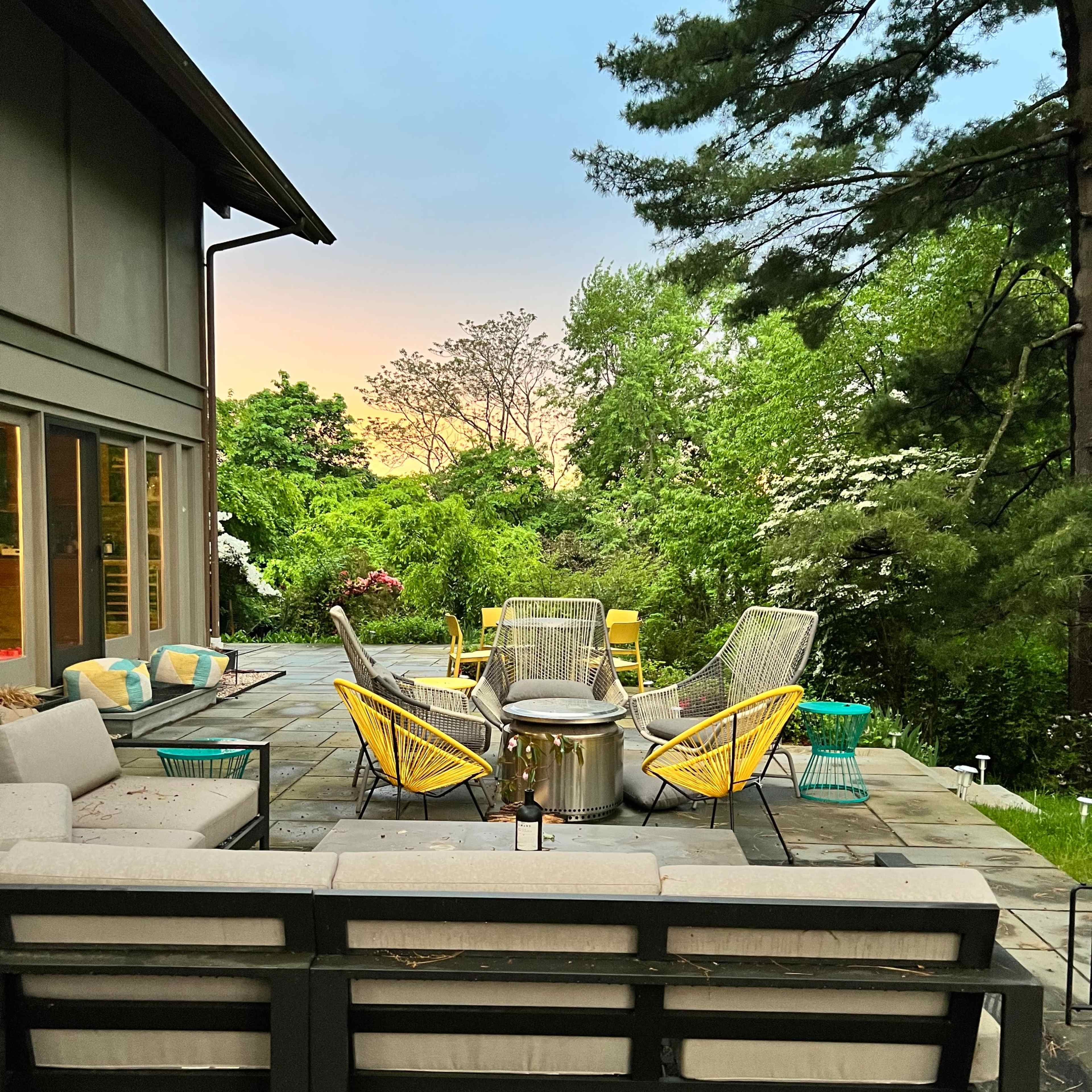 A stone patio features a circular fire pit surrounded by yellow and gray chairs and a sectional sofa, with green trees and a colorful sky in the background.