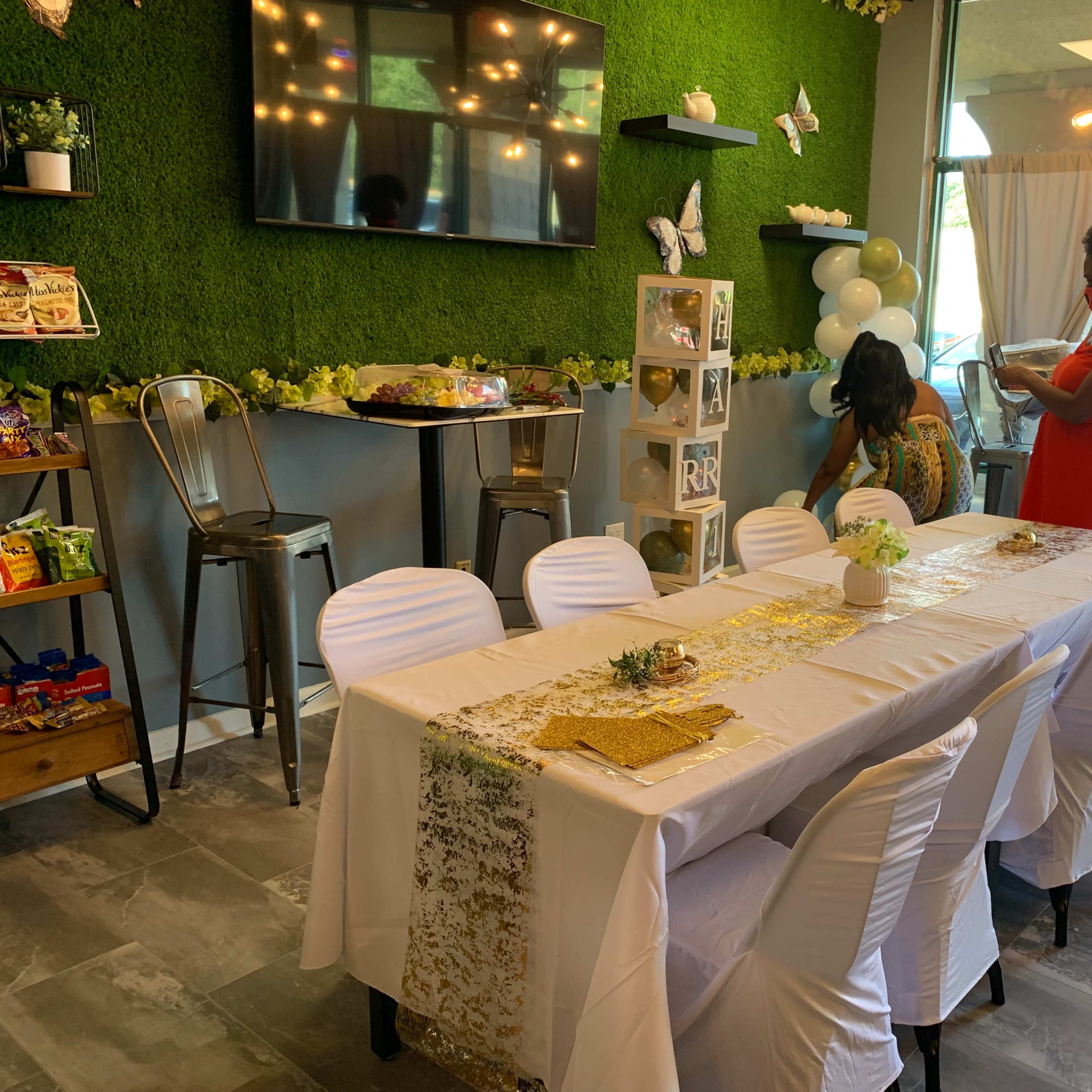 A decorated dining area features a long table set with a white tablecloth and golden accents, surrounded by chairs, while two women prepare for an event in a space adorned with greenery and butterfly decor.