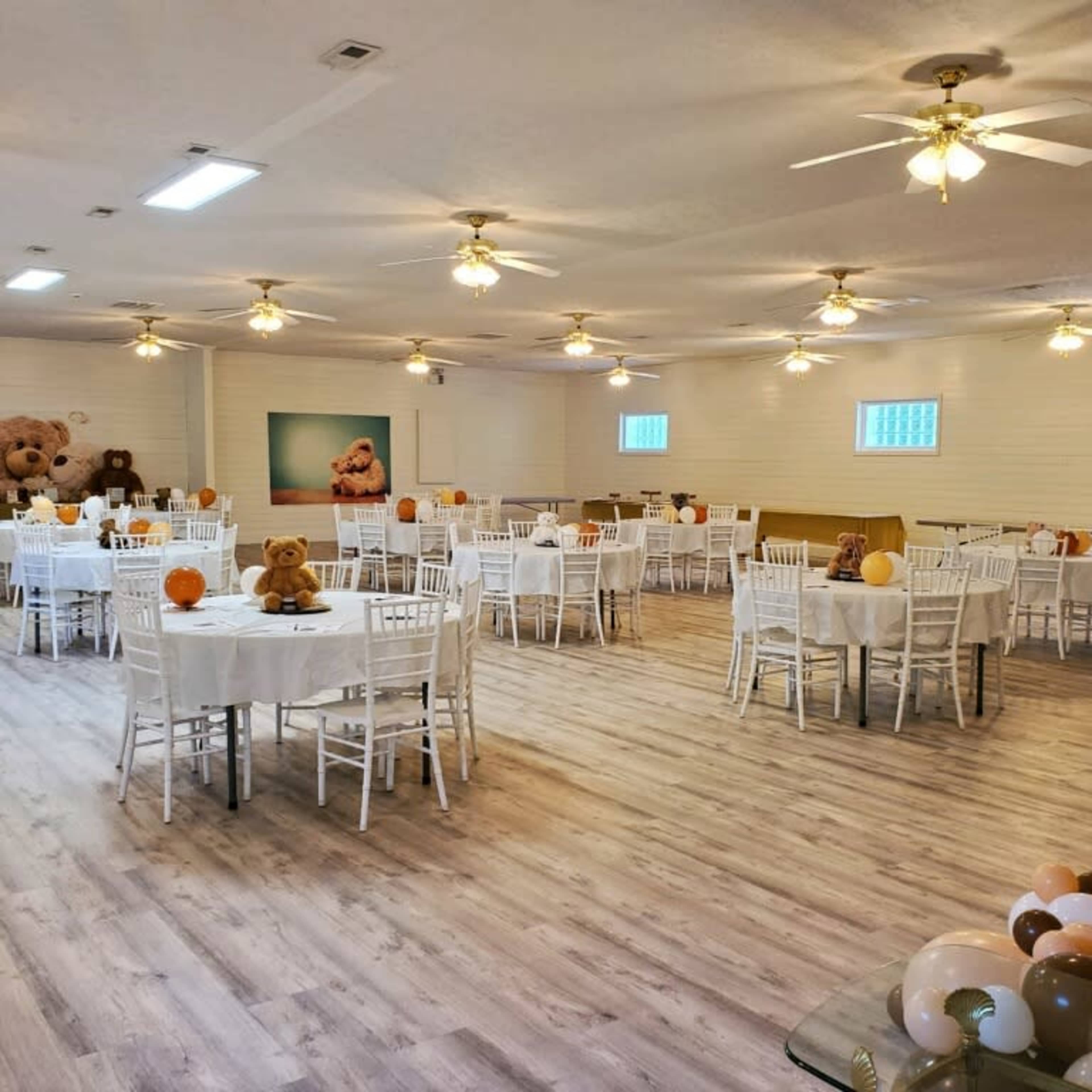 A spacious event room with white tables set for an occasion, surrounded by decorative teddy bears and balloons.