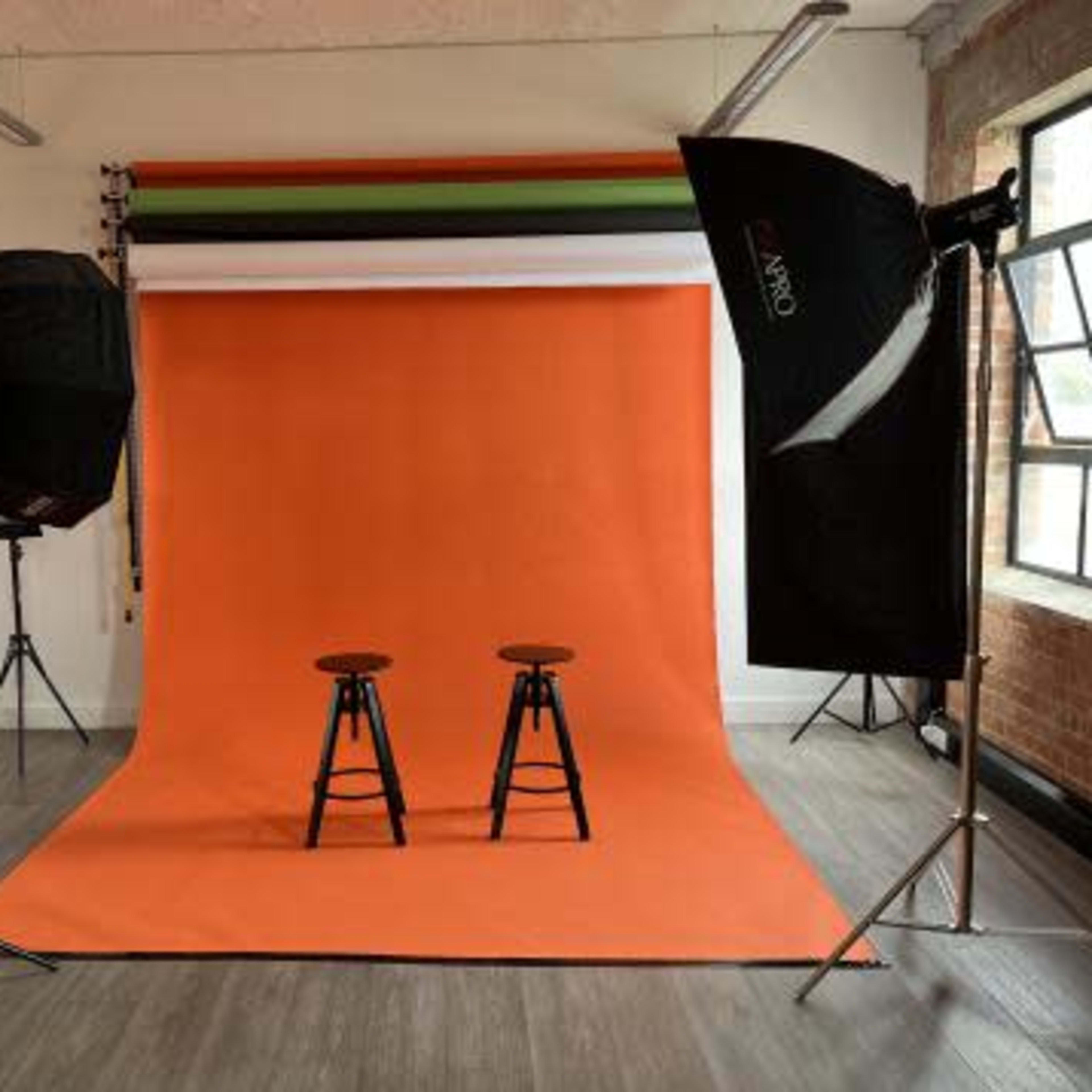 A photography studio with two wooden stools positioned in front of an orange backdrop, surrounded by lighting equipment.