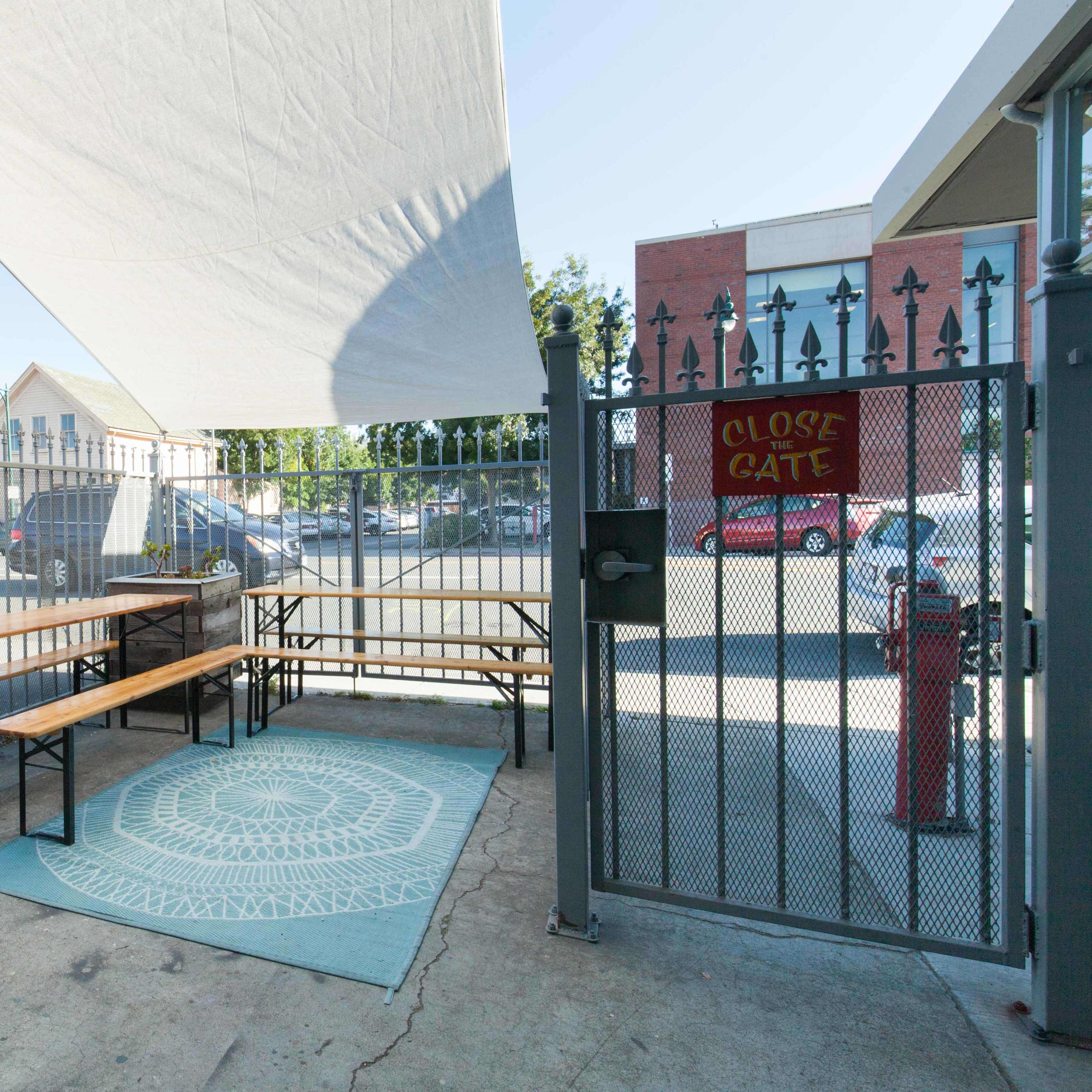 A shaded outdoor area features a wooden bench and table set on a patterned rug, enclosed by a fence with a closed gate sign.