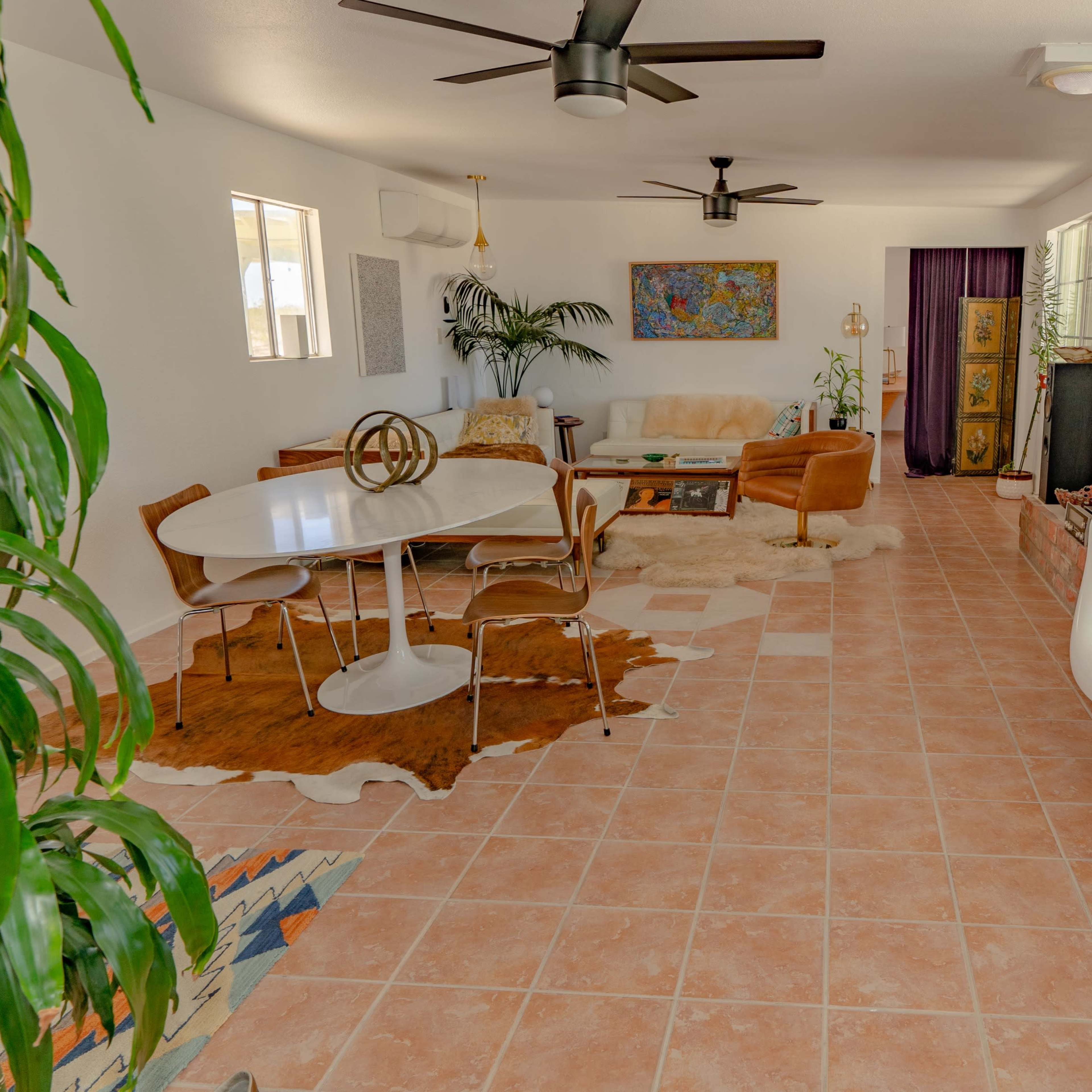 A spacious living area with tiled flooring, a round dining table, brown sofas, and large windows allowing natural light, surrounded by greenery.