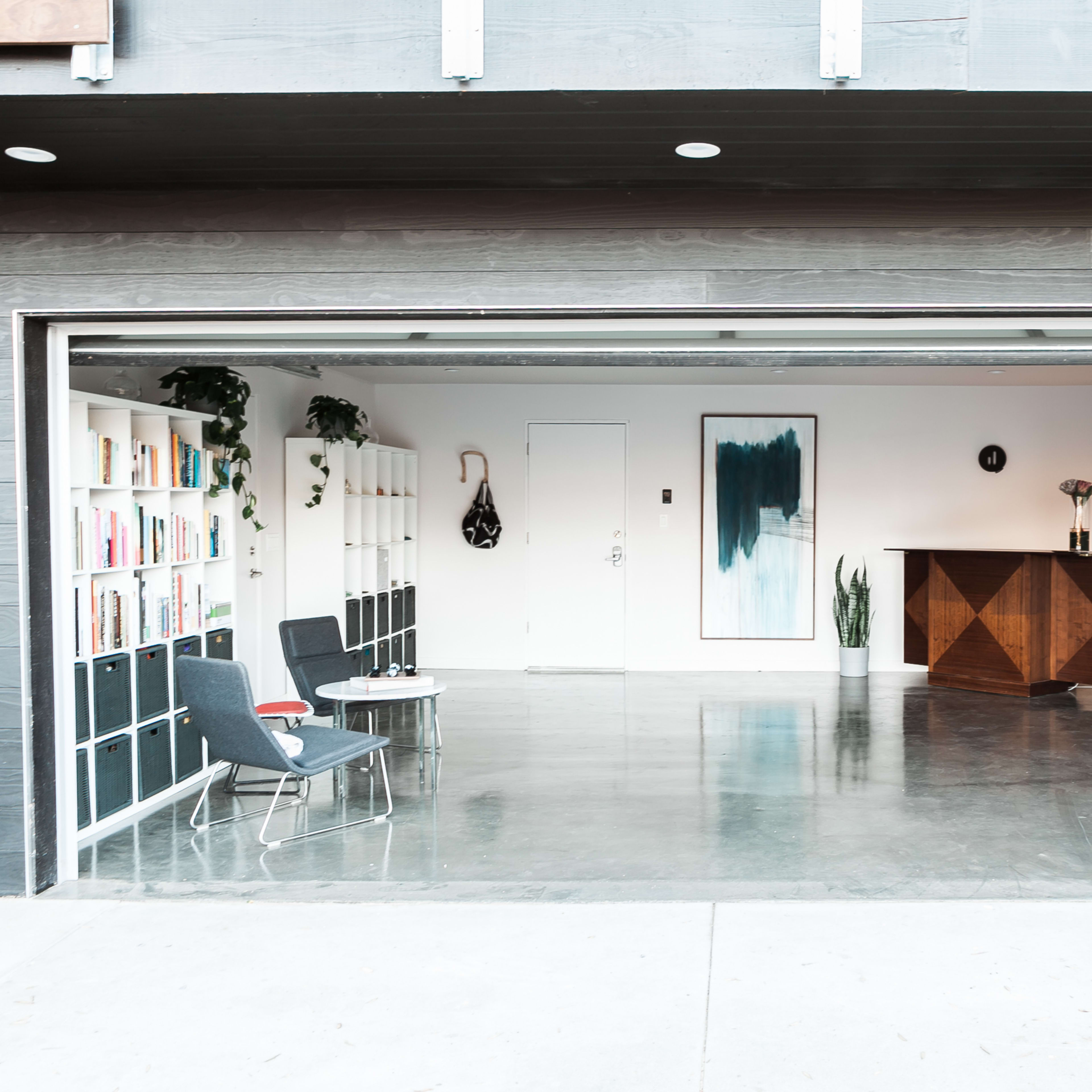 A spacious, modern indoor area with a large open garage door, featuring a bookshelf, a decorative table, and minimalist furniture.