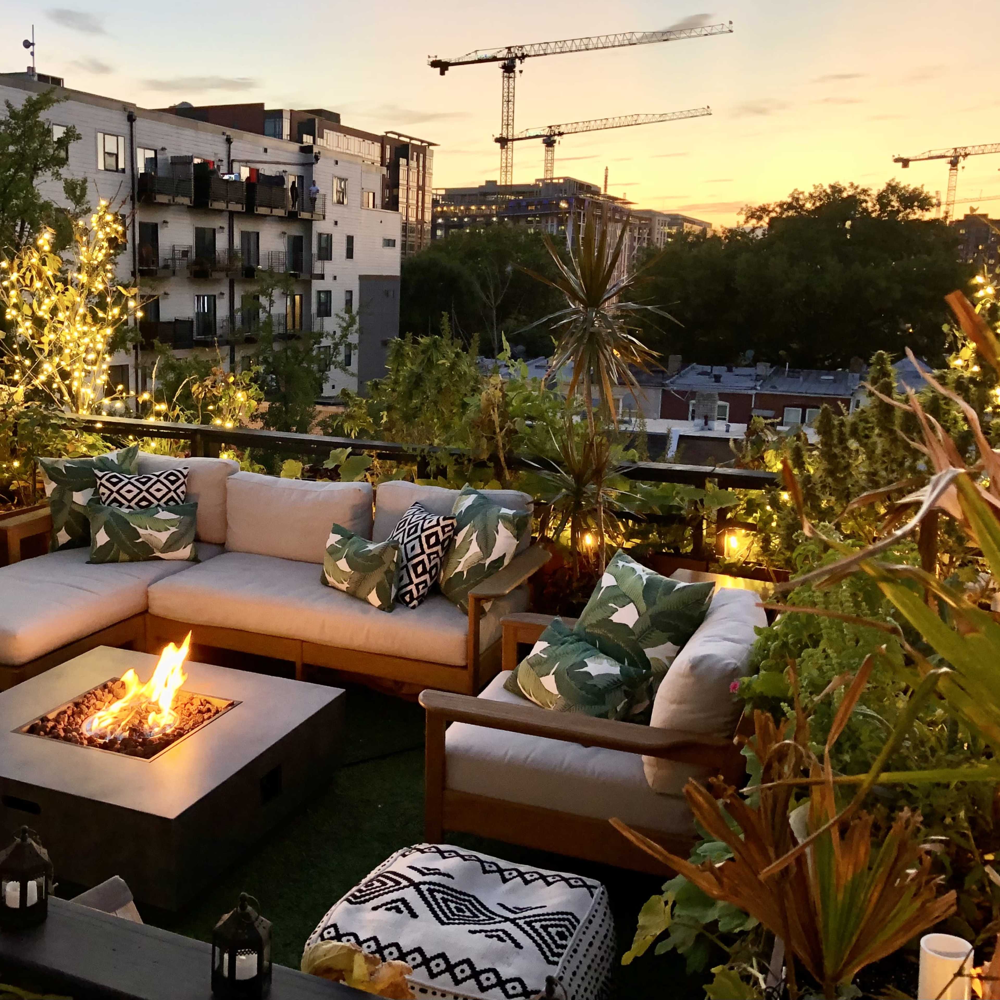 A rooftop patio with a sectional sofa, a fire pit, and potted plants, all illuminated by string lights at sunset.