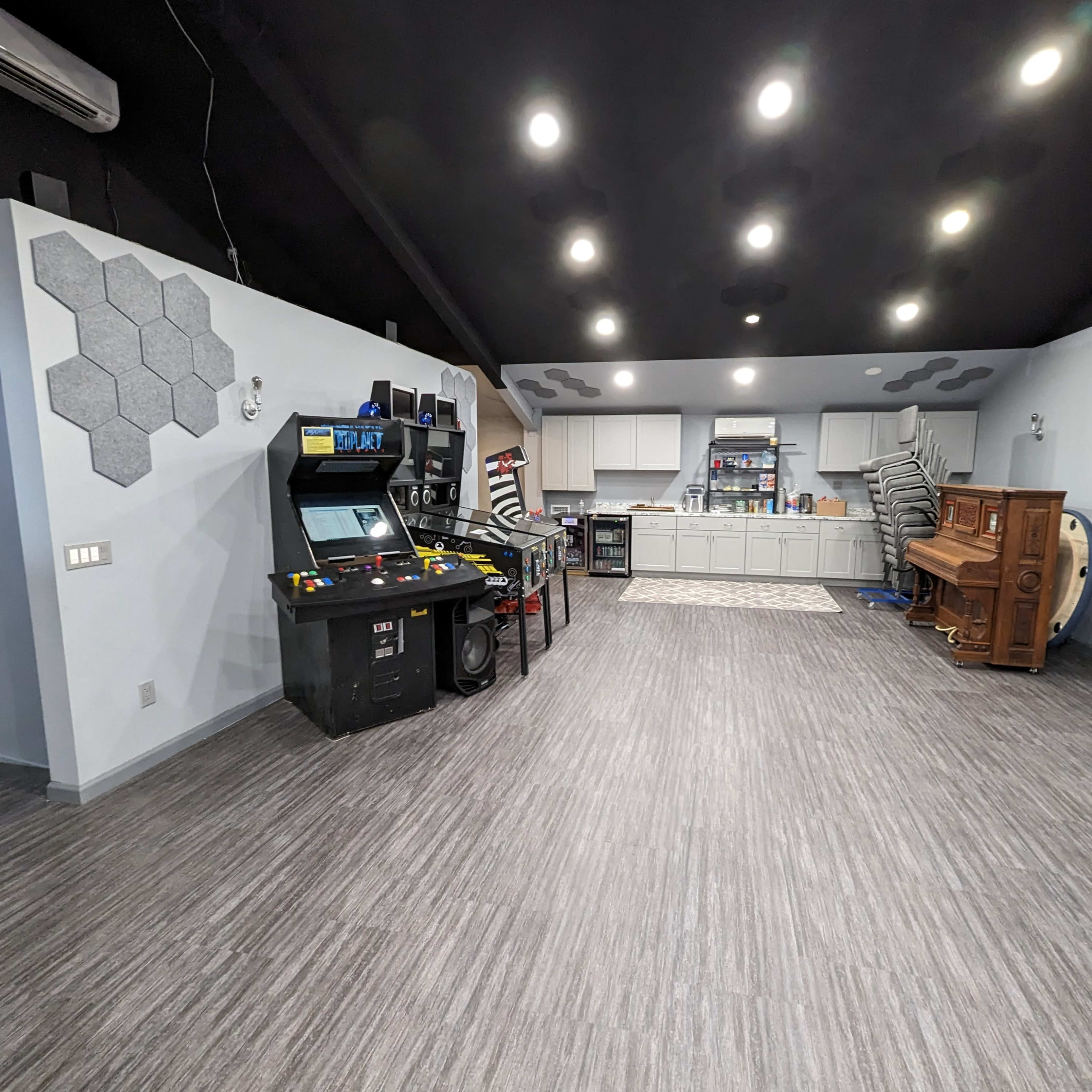 A spacious recreational room featuring a retro arcade machine, a stacked wooden piano, and a kitchenette area against a light gray wall with modern hexagonal decor.