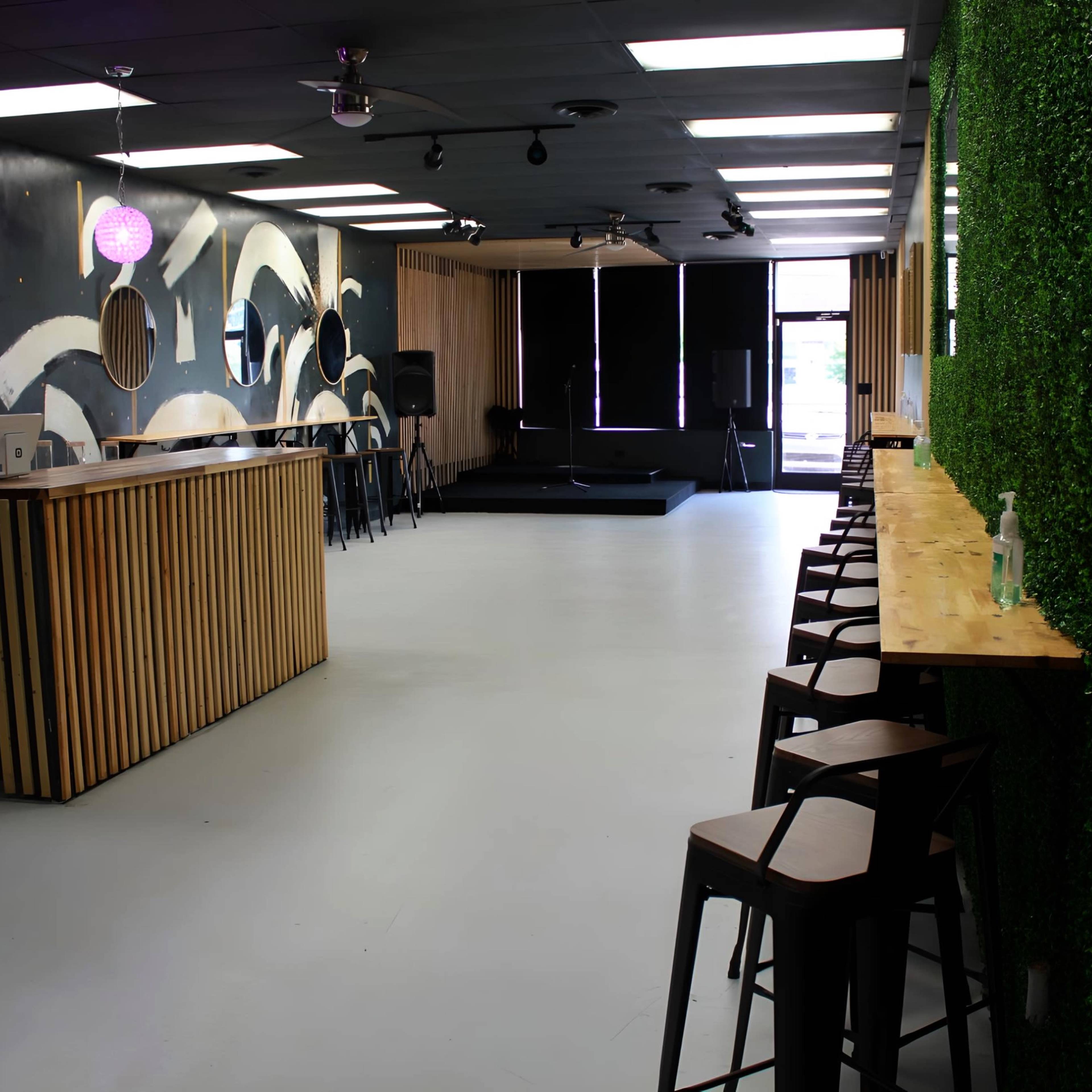 A spacious, modern interior of a bar with wooden accents, a long countertop, and green foliage on one wall, along with a small stage area at the back.