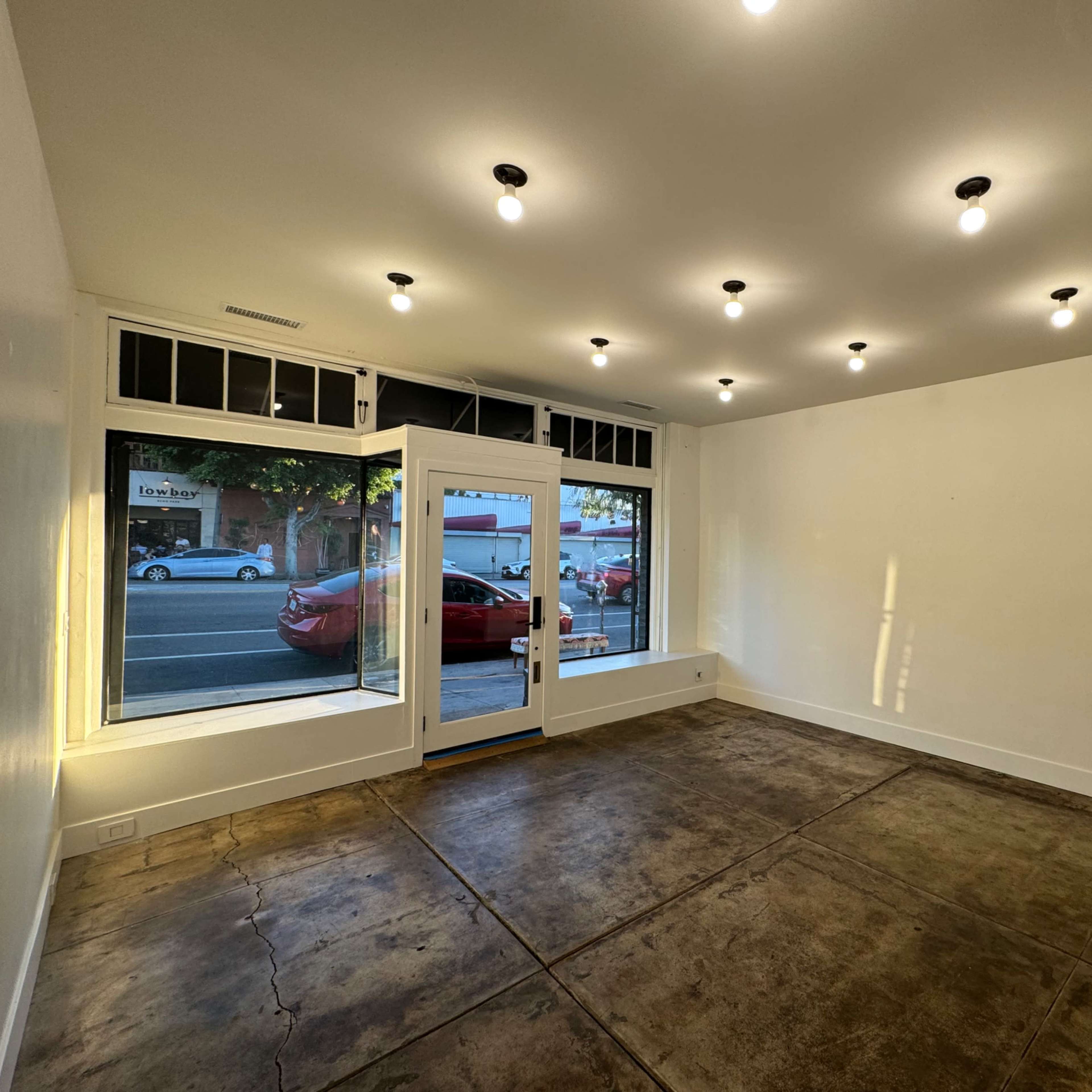 An empty room with concrete flooring, white walls, and multiple ceiling lights, featuring a large window facing a street.