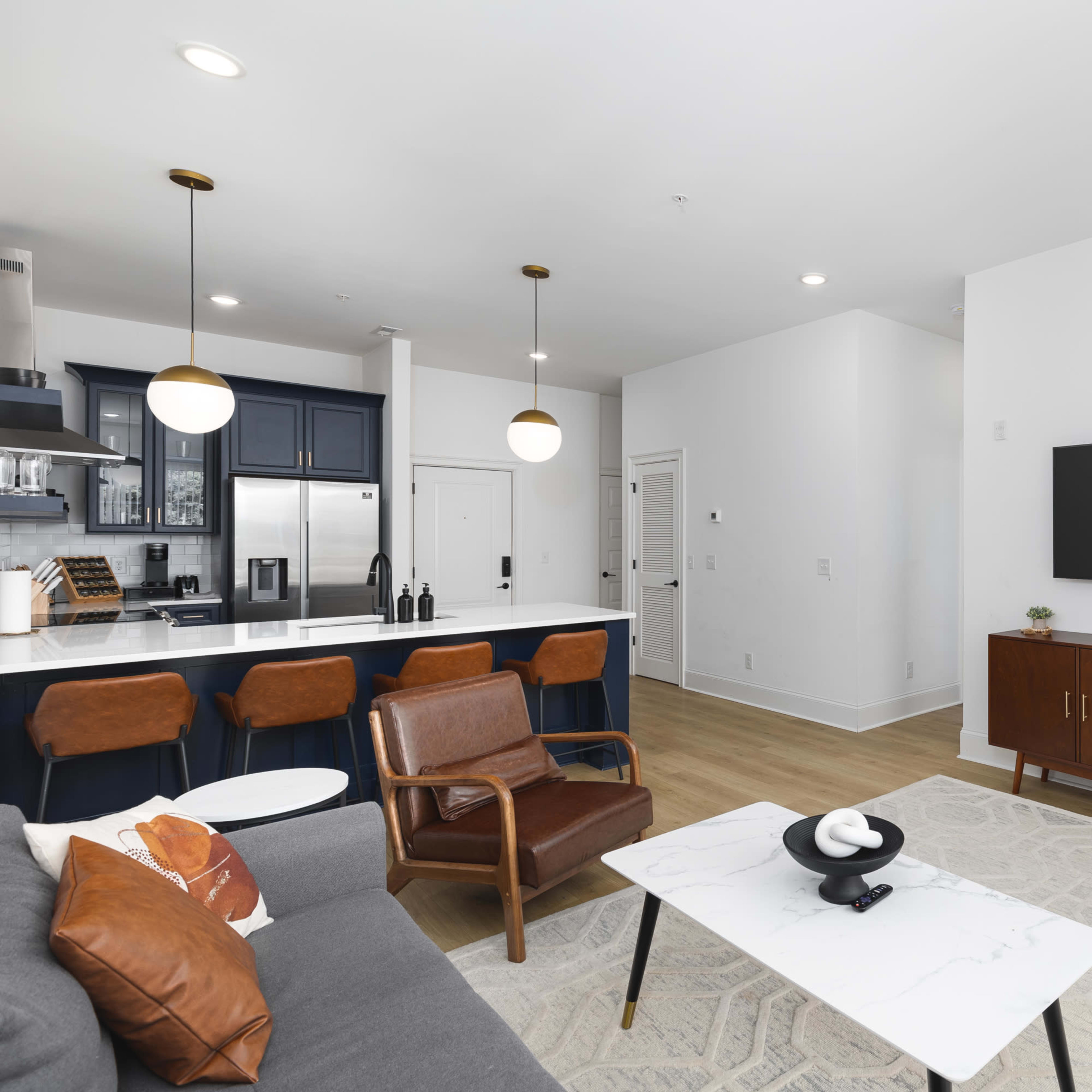A modern living room features a gray sofa, a coffee table, a wooden sideboard, and a wall-mounted television, with a kitchen visible in the background.