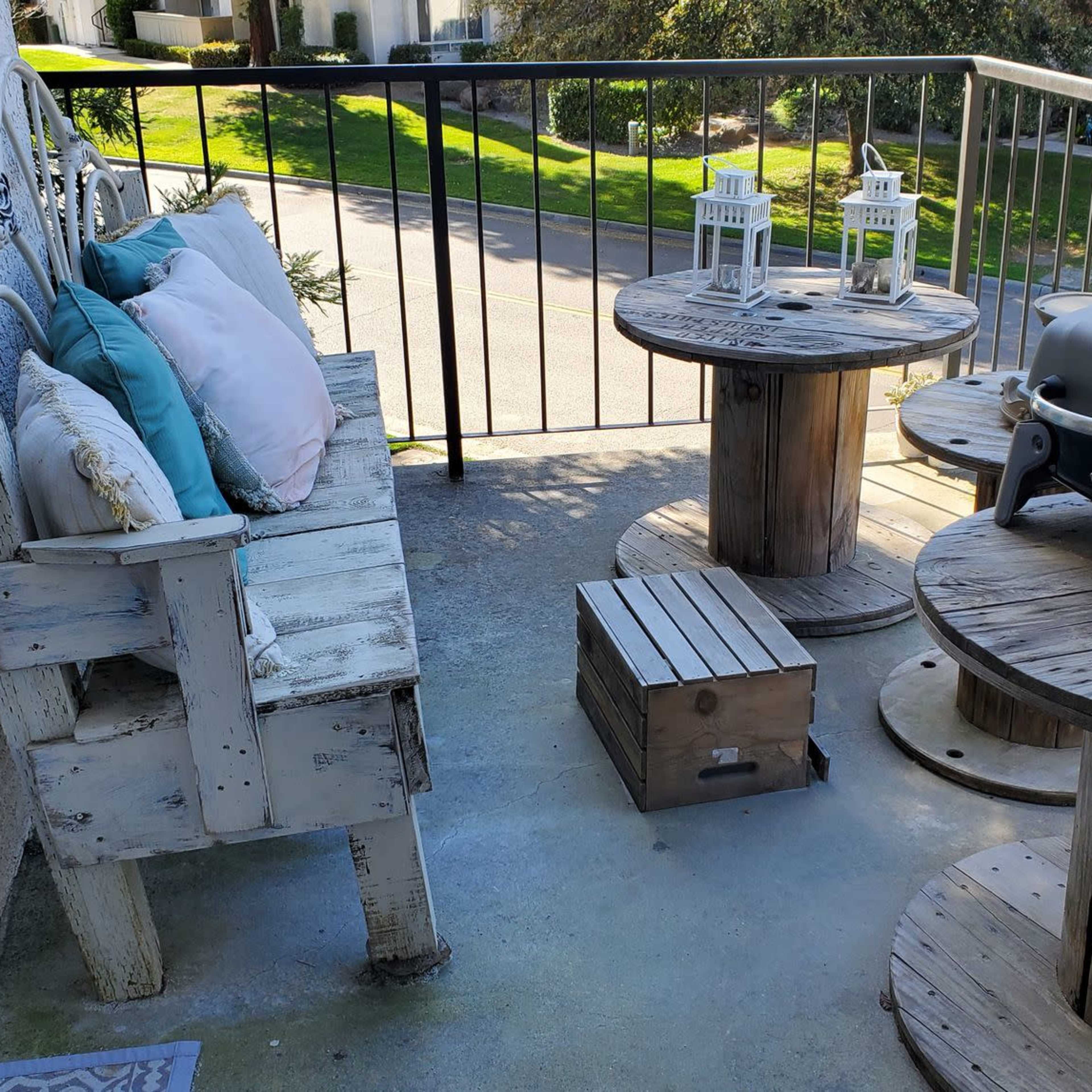 A balcony with a wooden bench, two round tables made from pallets, a portable grill, and decorative lanterns.