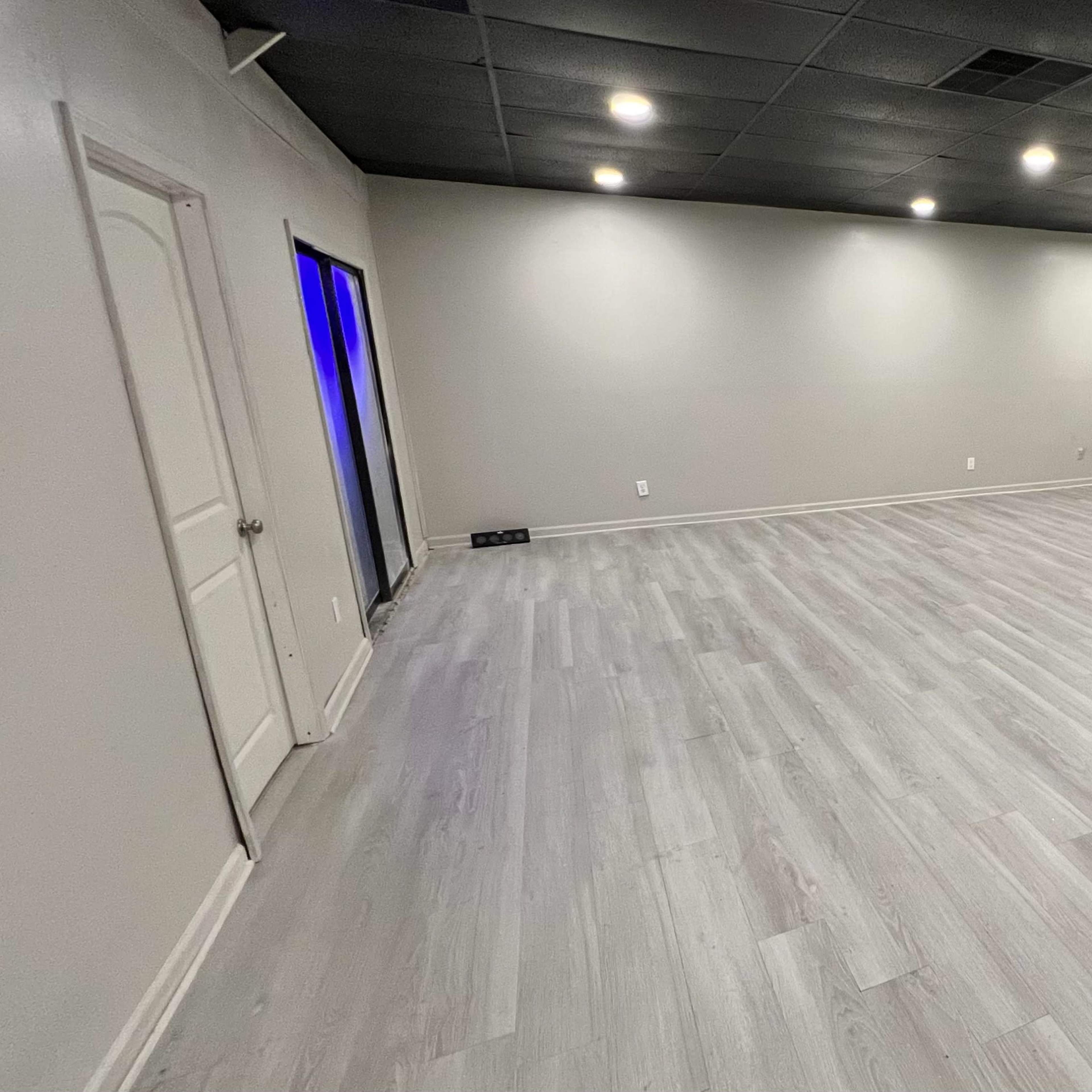 A spacious, empty room with gray walls and light-colored wooden flooring, featuring several overhead lights and a door on the left.