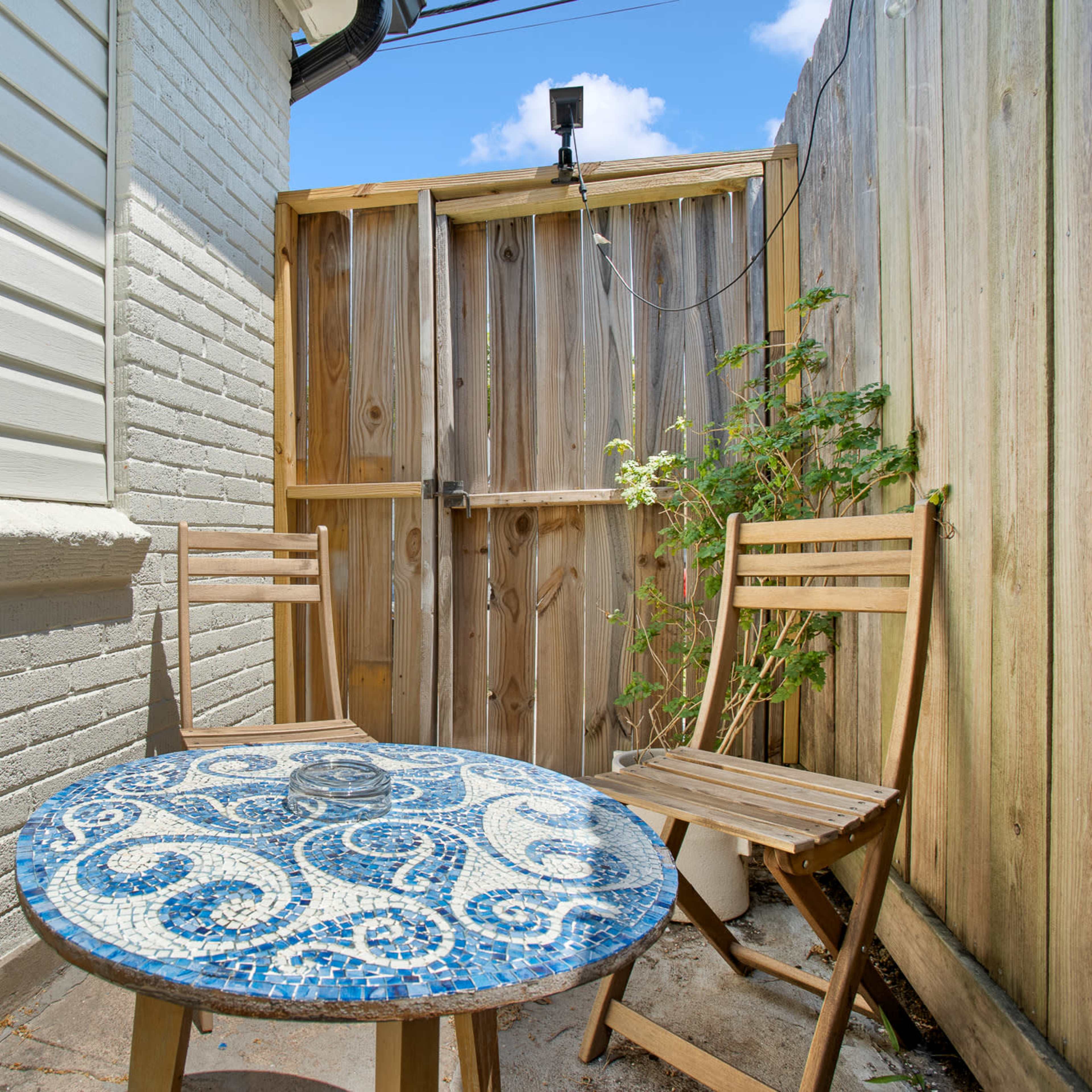 A small outdoor seating area features a mosaic-topped table and two wooden chairs against a wooden fence.