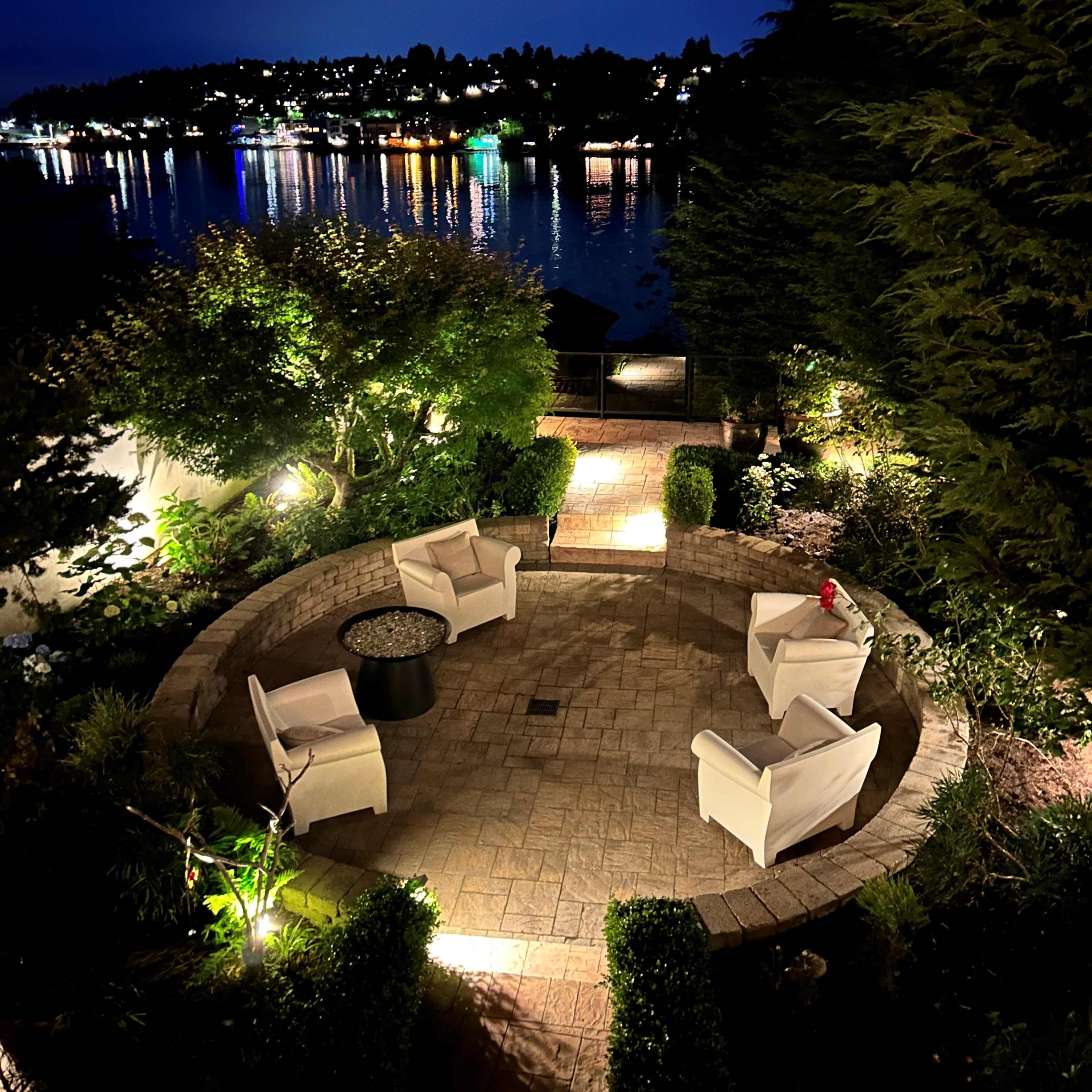 A circular stone patio with four white chairs and a small table overlooks a tranquil lake illuminated by distant lights at night.