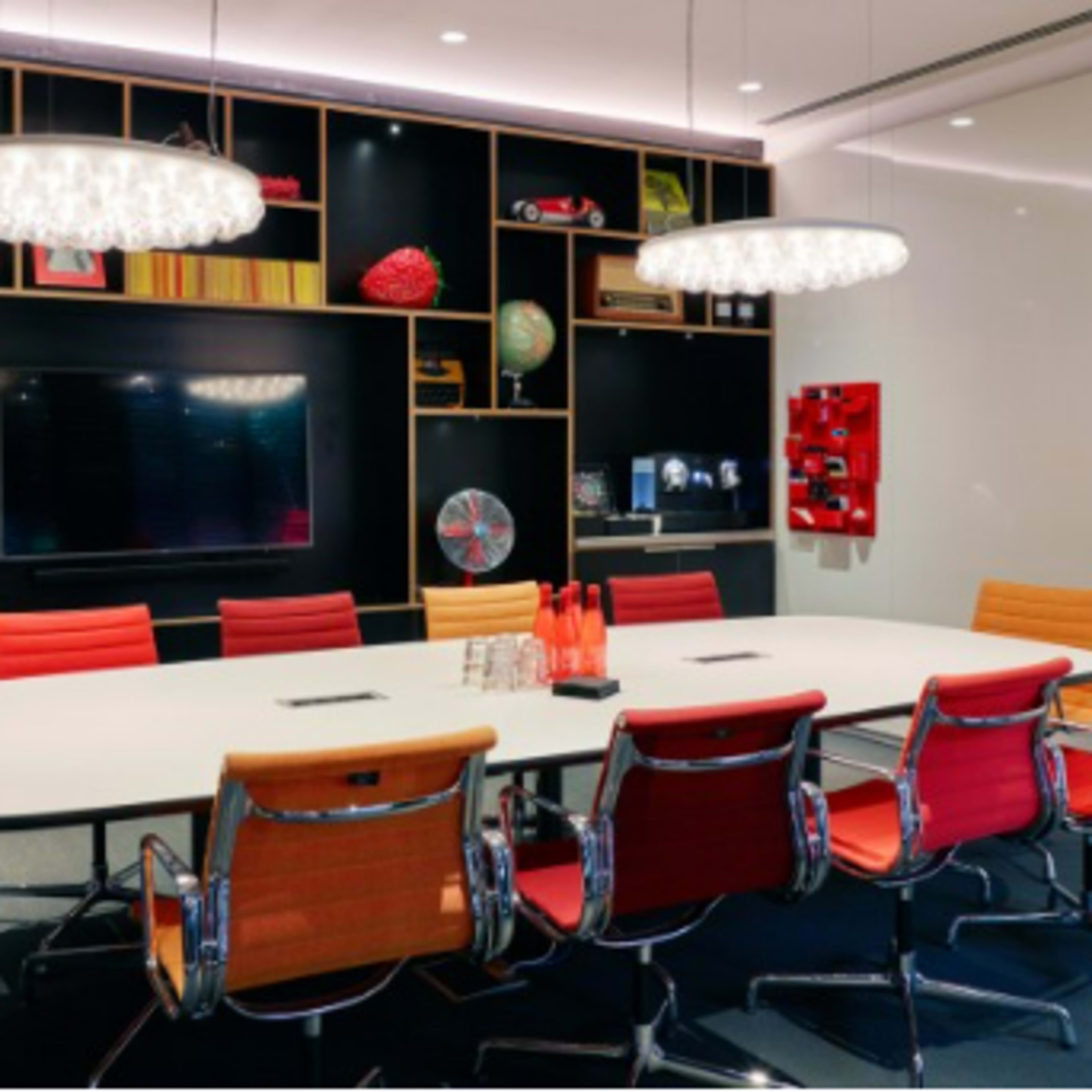 A modern conference room with a large white table surrounded by red and orange chairs, featuring decorative shelves with various items and a television mounted on the wall.