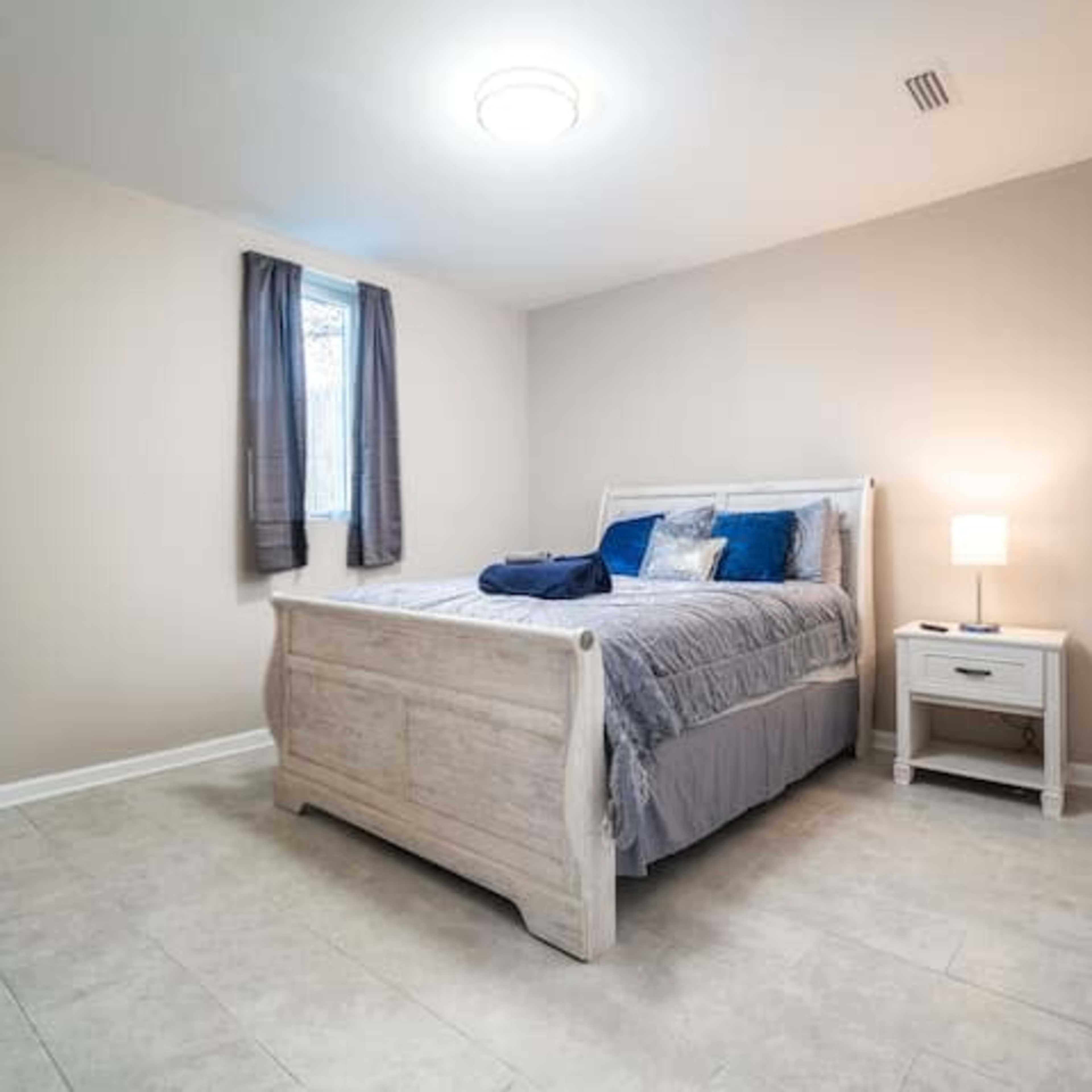 A simple bedroom featuring a bed with a gray and blue bedding set, a nightstand with a lamp, a television, and light-colored walls and flooring.