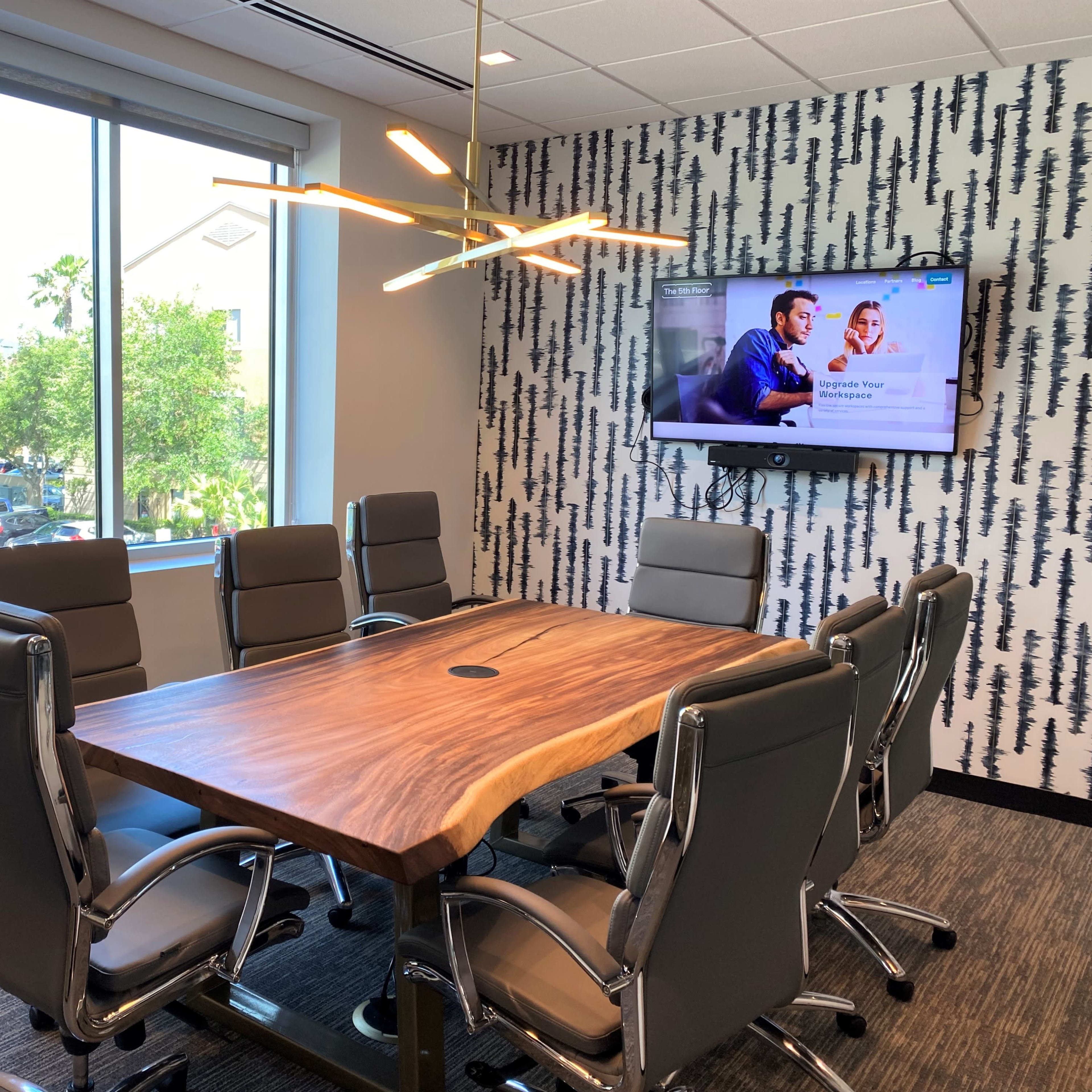 A modern conference room features a large wooden table surrounded by gray office chairs, with a wall-mounted TV displaying a presentation and large windows offering a view of palm trees outside.
