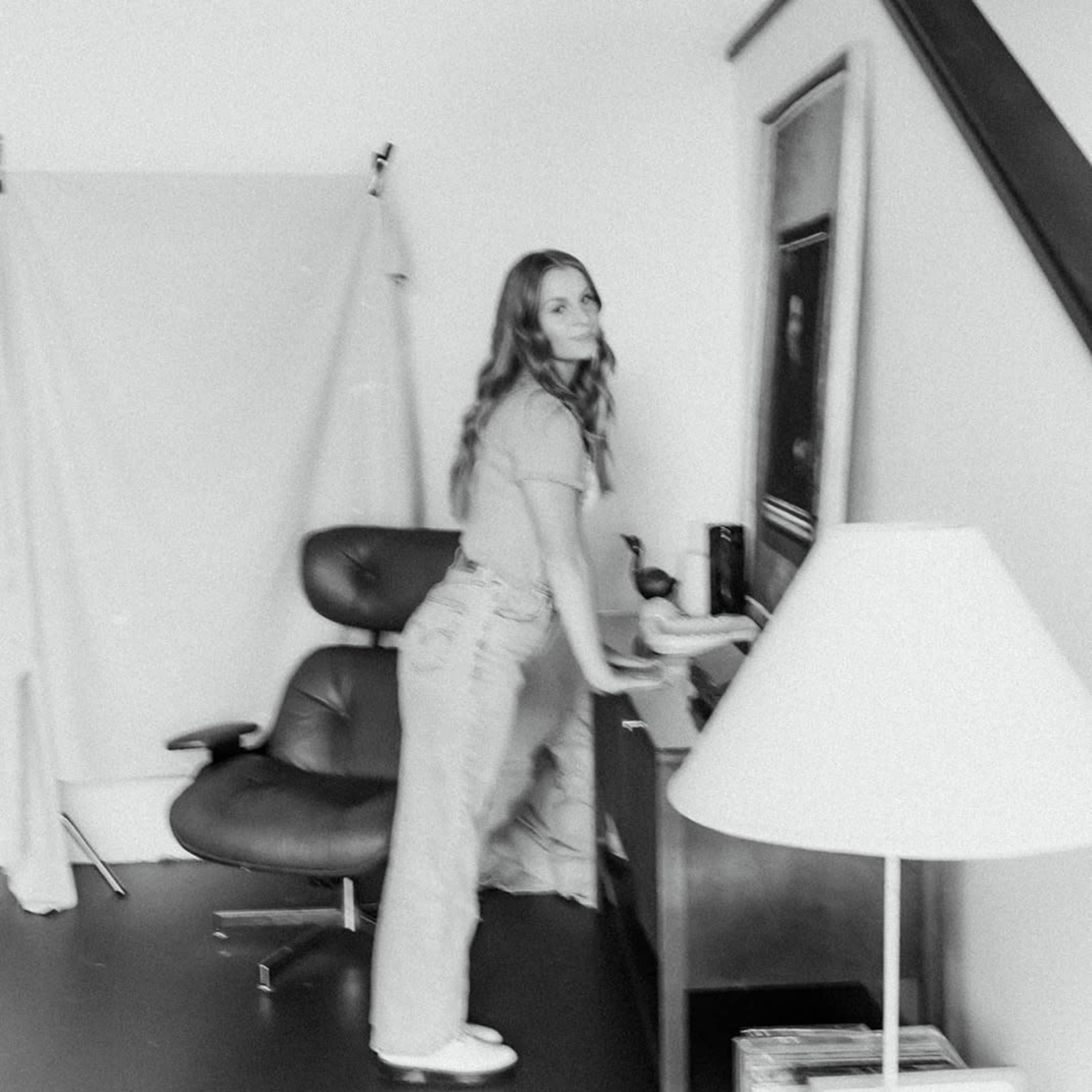 A woman stands at a desk in a room with a chair and a lamp, with a backdrop hanging nearby.