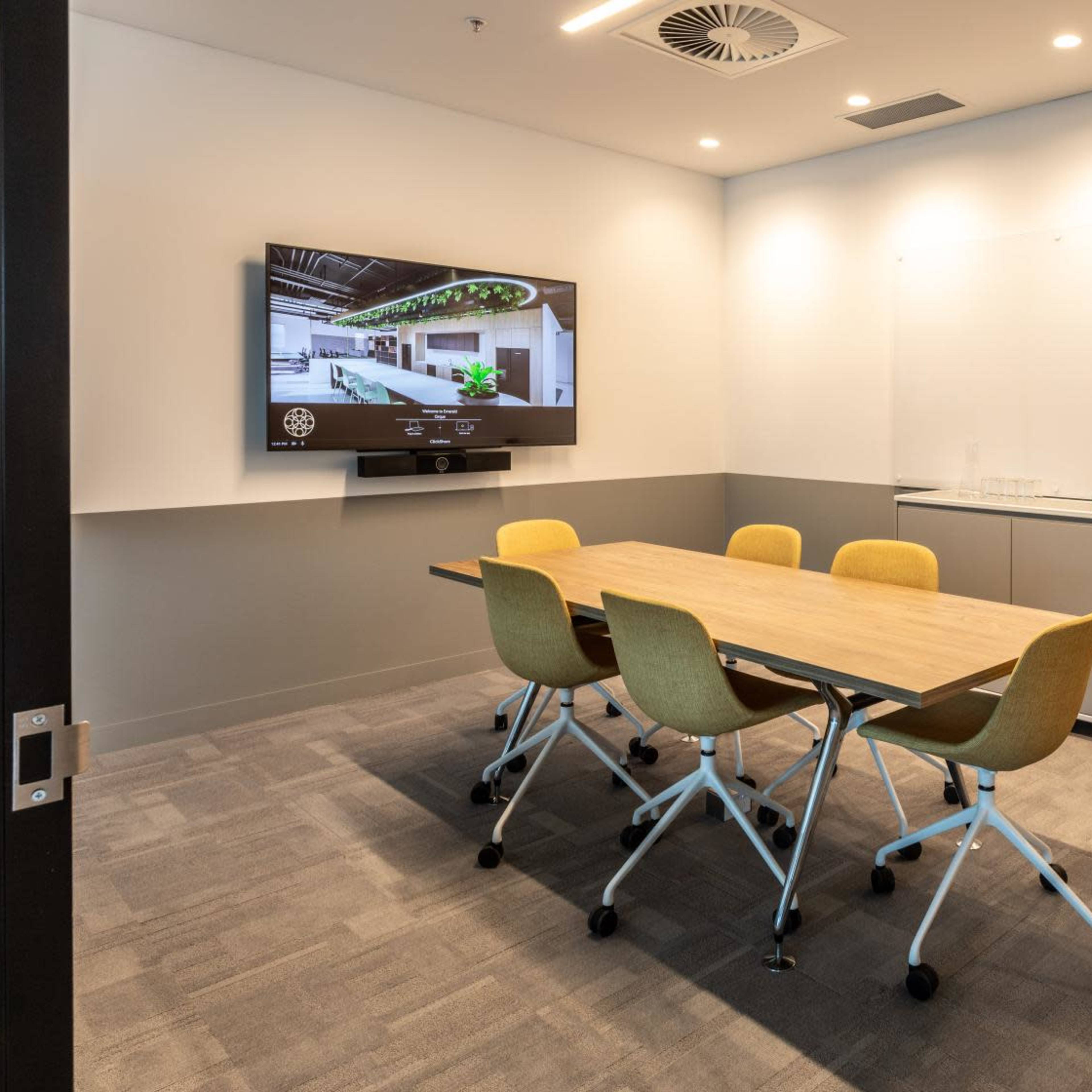 A small conference room features a wooden table surrounded by yellow chairs, a wall-mounted television, and a whiteboard.